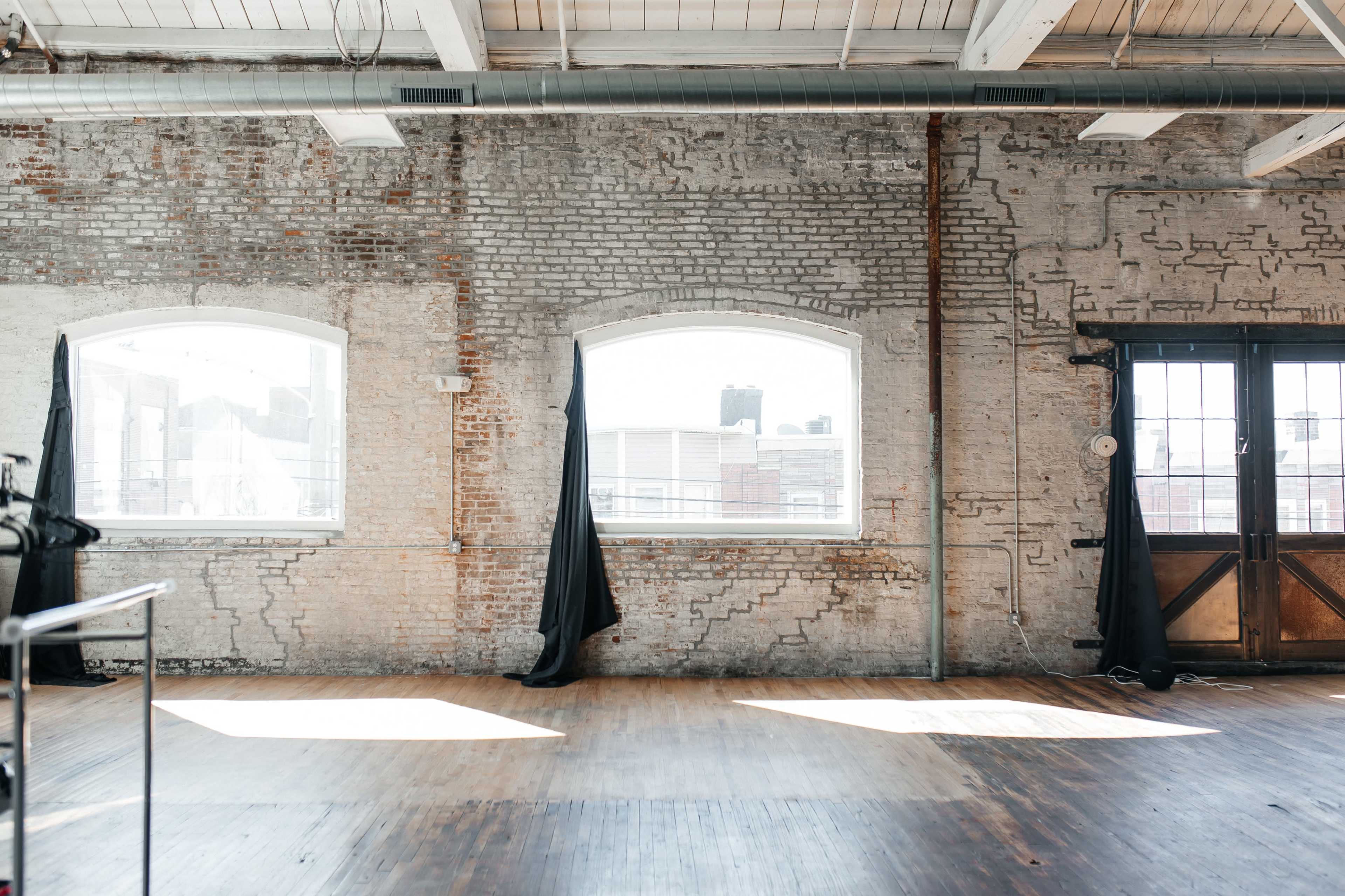 An industrial-style room with exposed brick walls, two large arched windows, and black curtains hanging on either side.