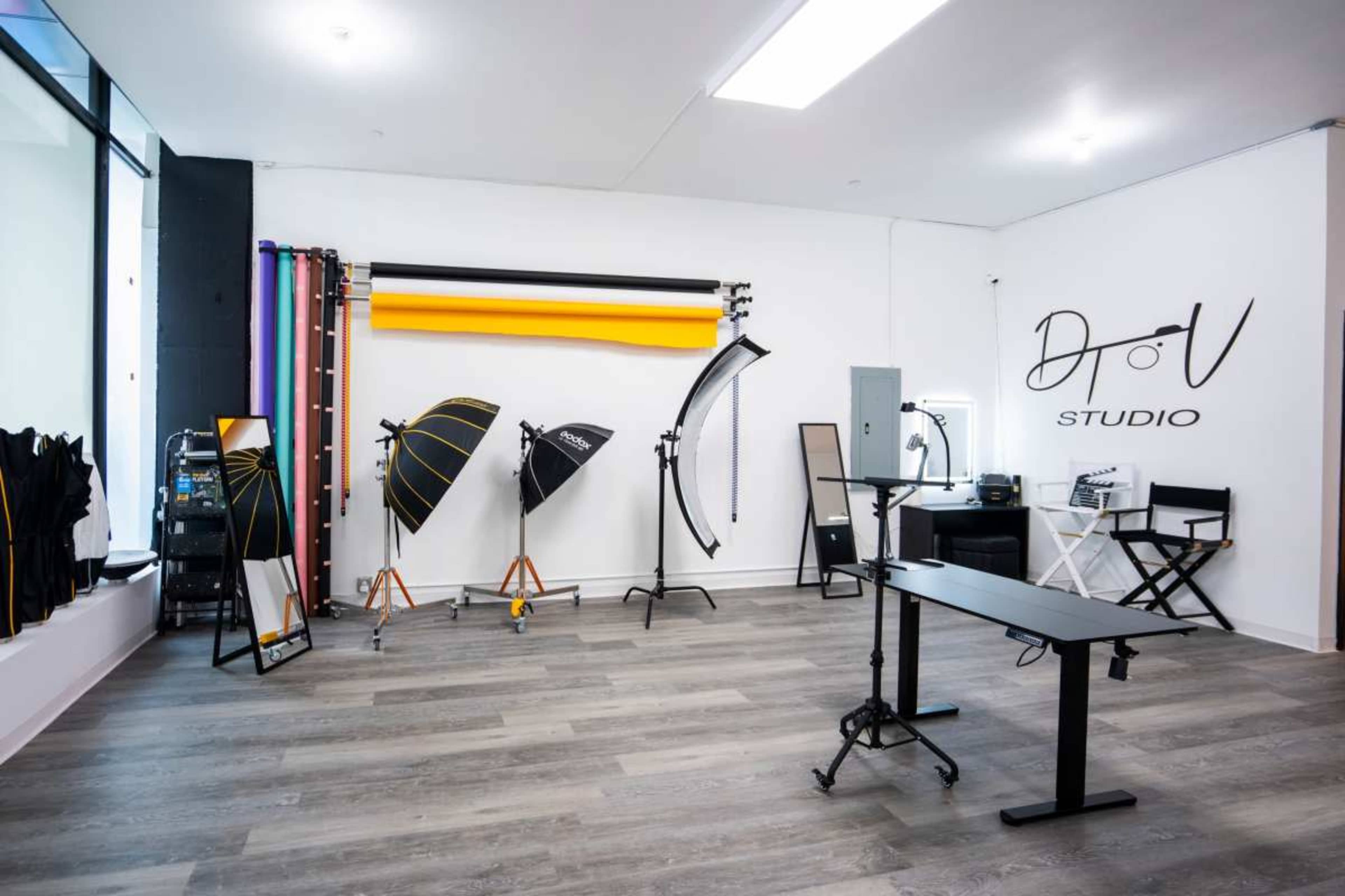 The image shows a photography studio featuring various lighting equipment, backdrops, and a workspace with a chair and table.