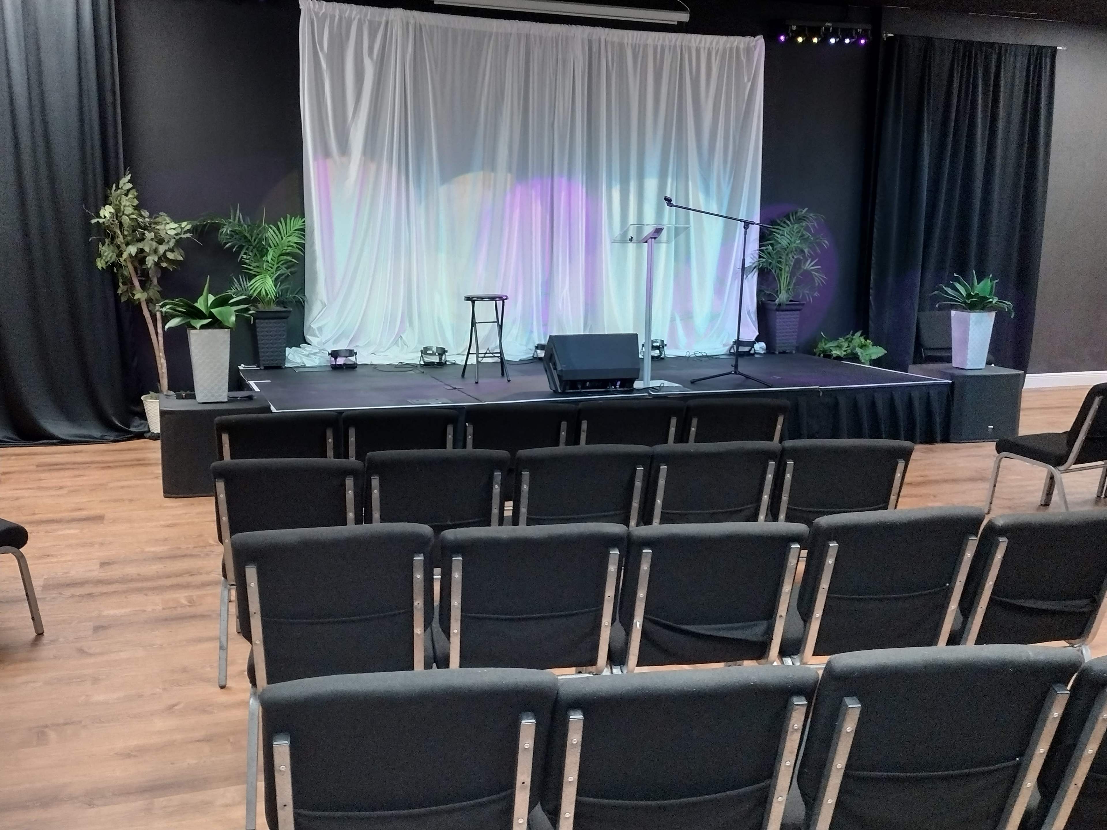 A stage with a microphone, music stand, and chairs arranged in front of it, set against a backdrop of a white curtain and plants.