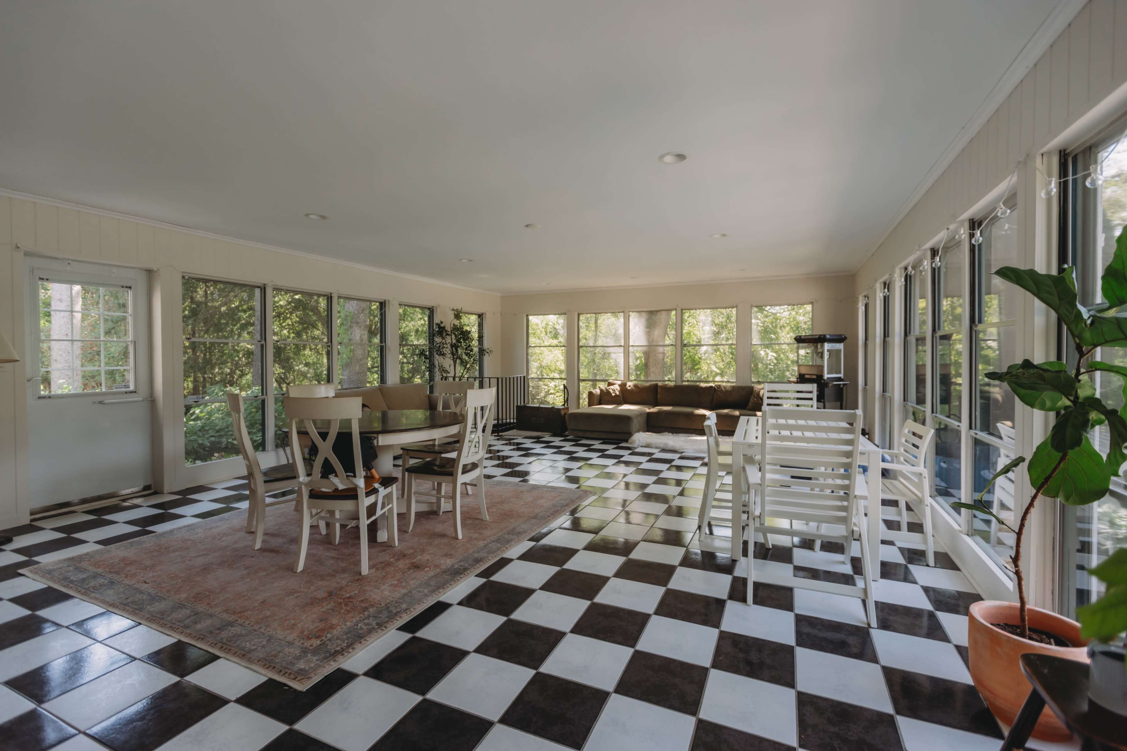 The image shows a bright sunroom featuring a checkered black and white tiled floor, a dining area with chairs and a table, and a seating area with a couch and large windows.