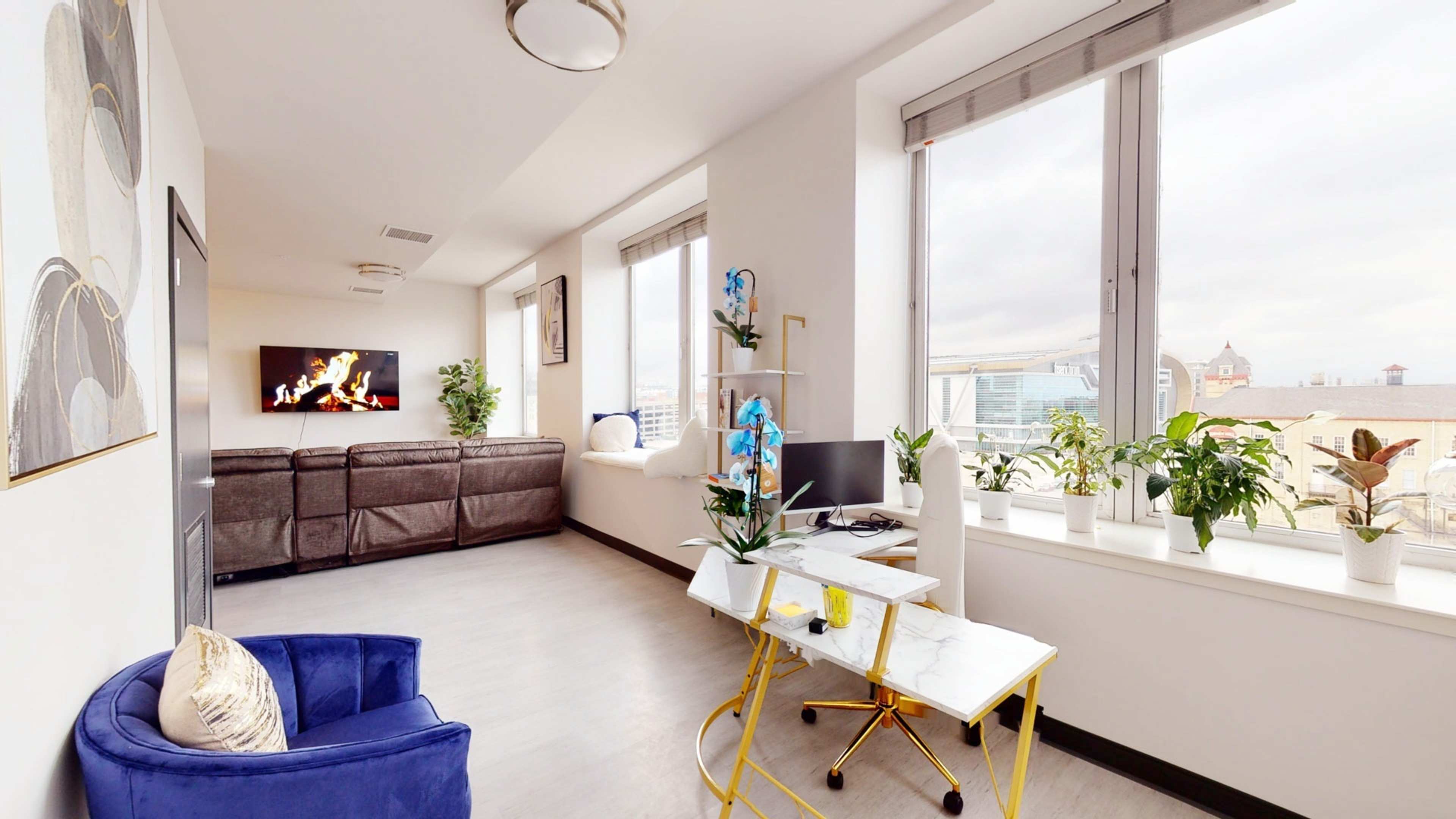 A modern living room features a blue chair, a small desk with plants, and large windows overlooking the city.