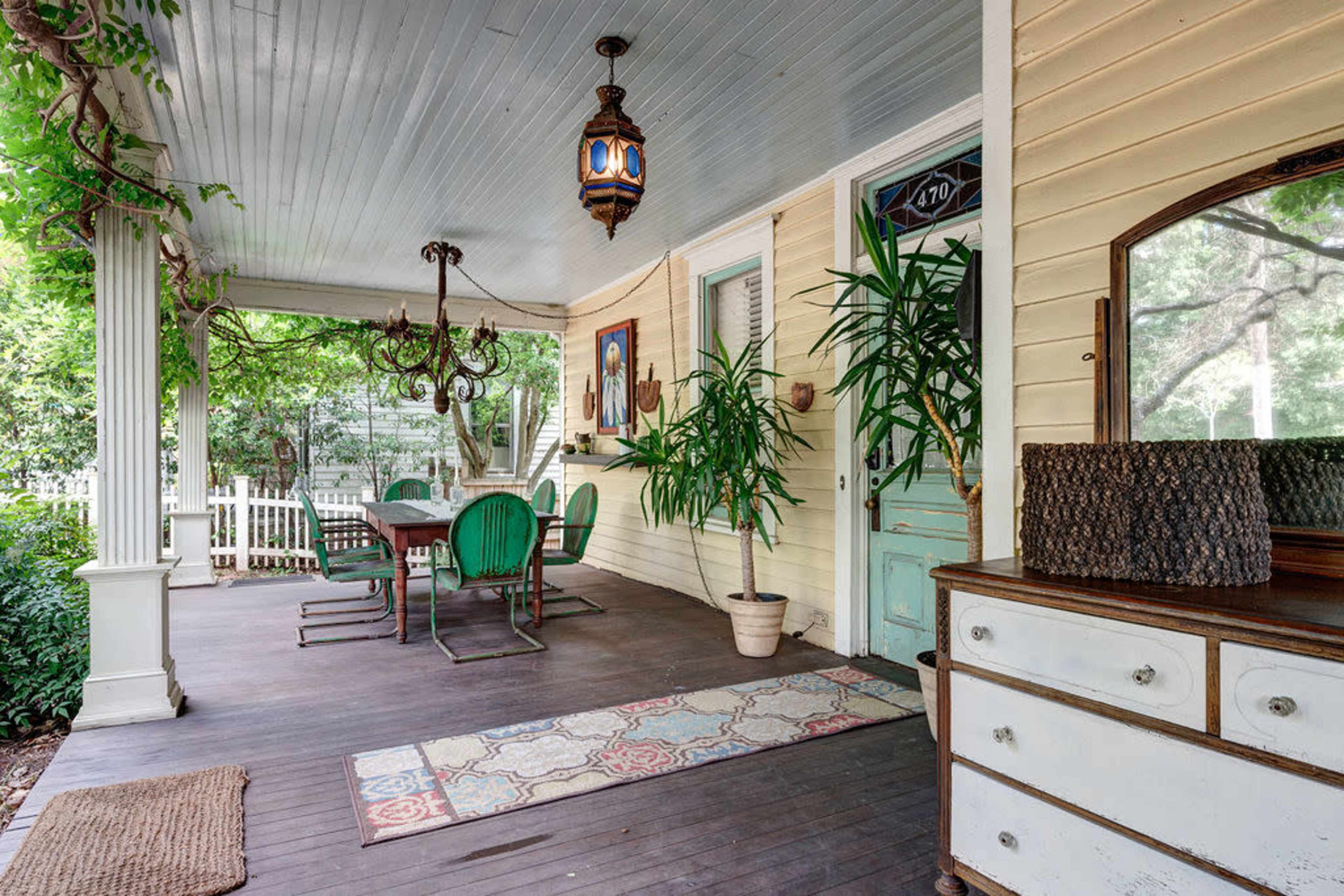 The image depicts a covered porch featuring a wooden table with green chairs, potted plants, and a vintage chest of drawers against a wall.