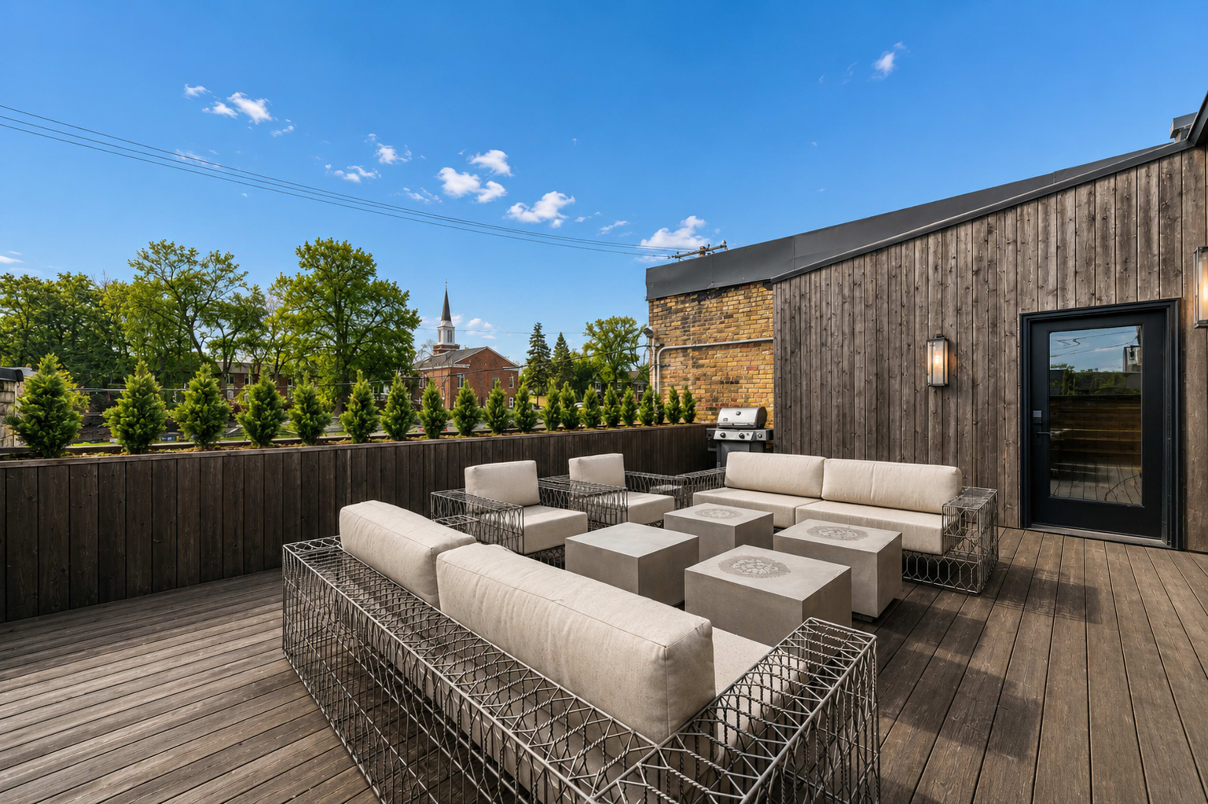 The image shows a rooftop deck featuring modern seating arrangements made of light-colored cushions and stone tables, surrounded by greenery and a view of a church steeple in the distance.