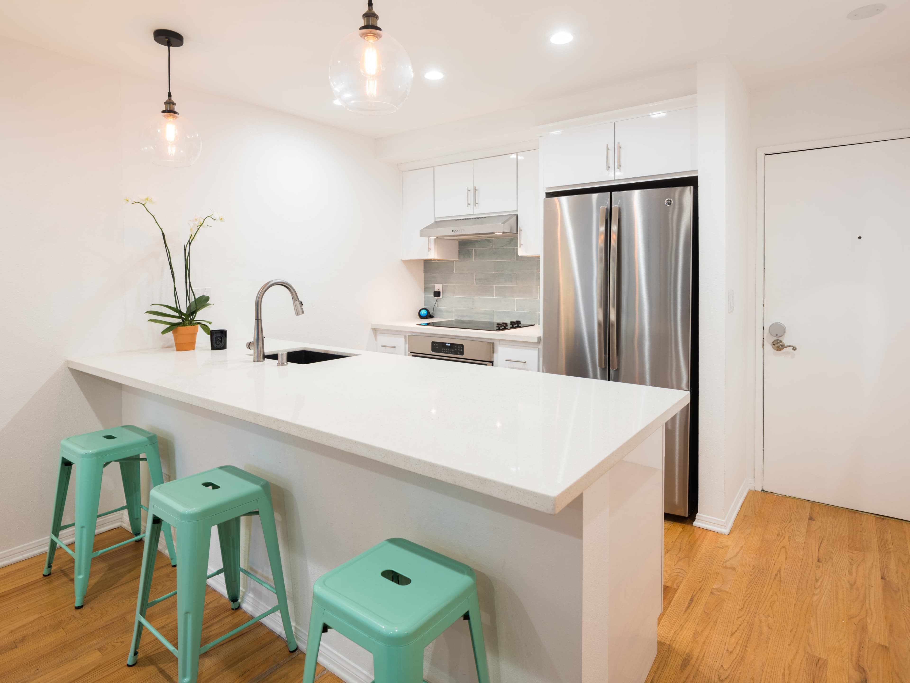The image shows a modern kitchen with a white countertop, stainless steel appliances, and three teal stools at the island.