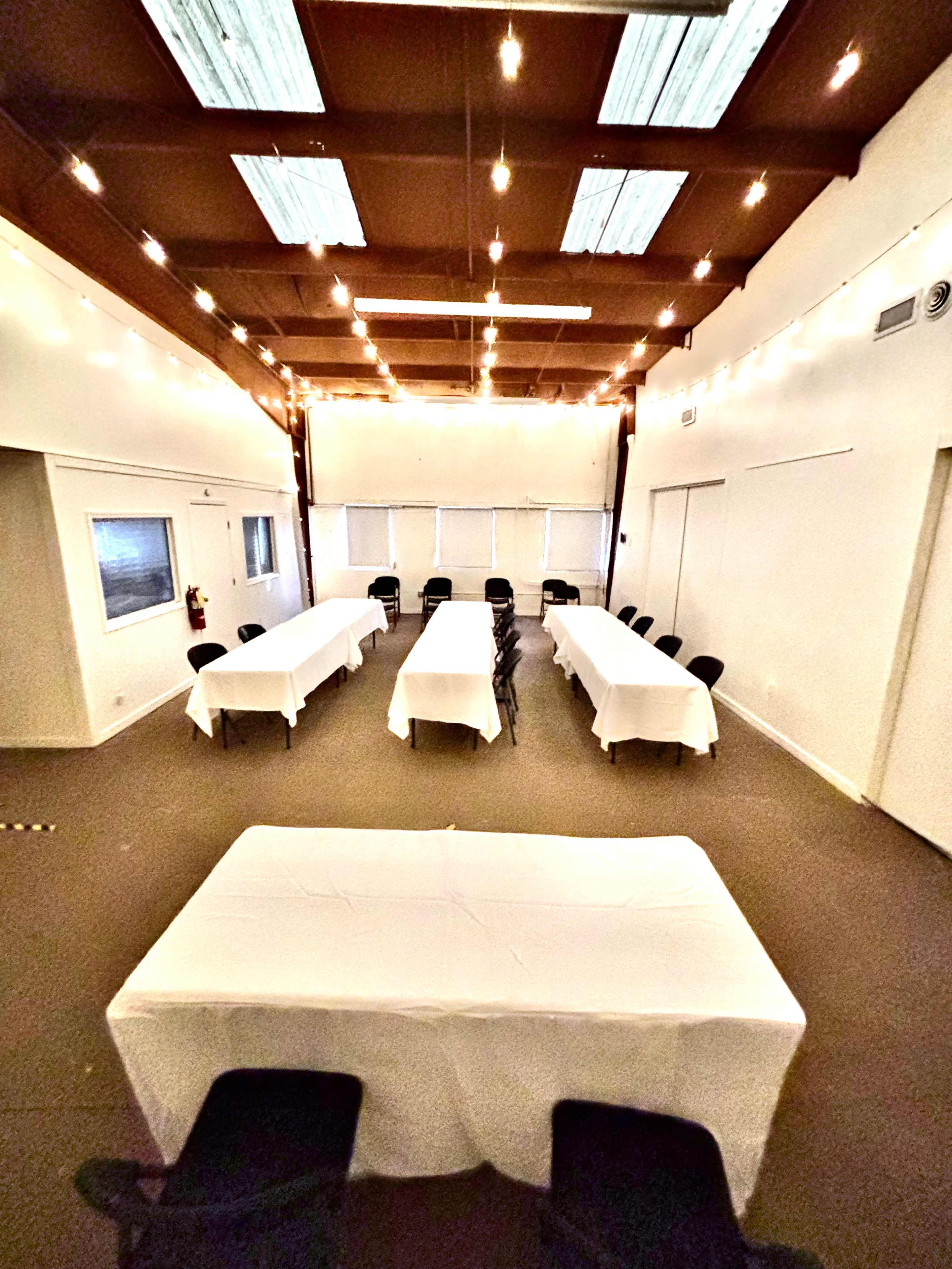 The image shows a spacious room set up with multiple tables covered in white tablecloths arranged in an open layout, illuminated by string lights overhead.