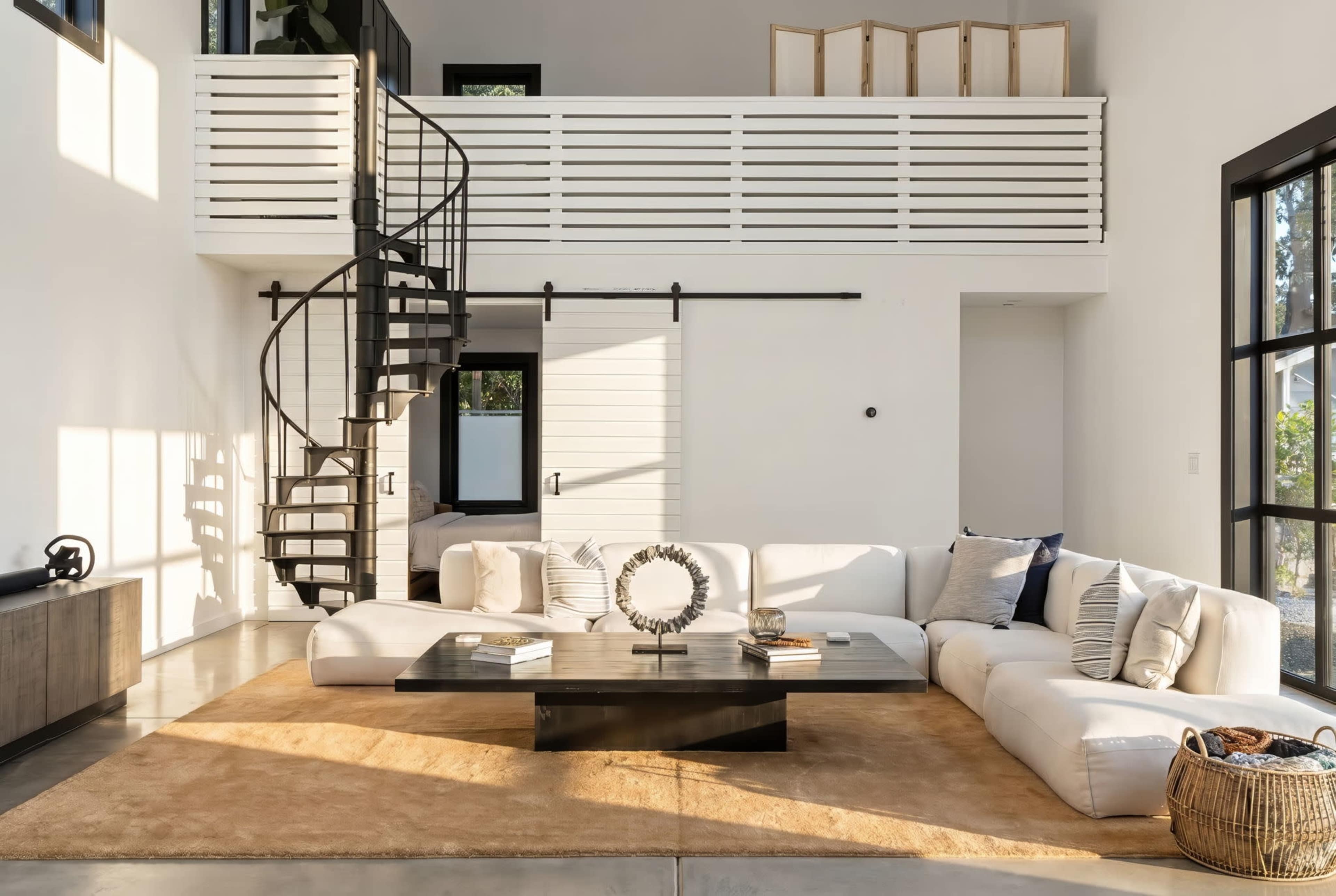 The image shows a modern living room with a spiral staircase, a large sectional sofa, a coffee table in the center, and large windows allowing natural light to fill the space.