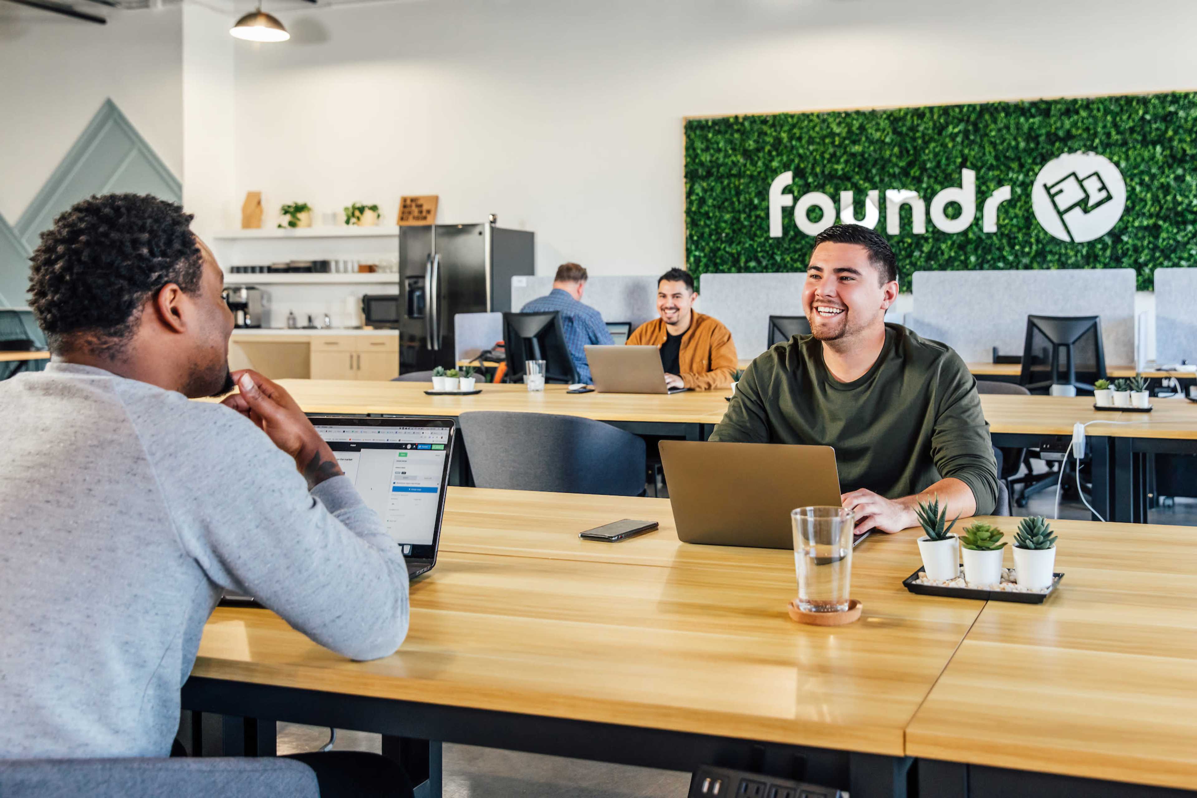 Two men are seated at a wooden table in a modern office space, with one smiling at the other while using laptops, and a green wall featuring the word "foundr" in the background.