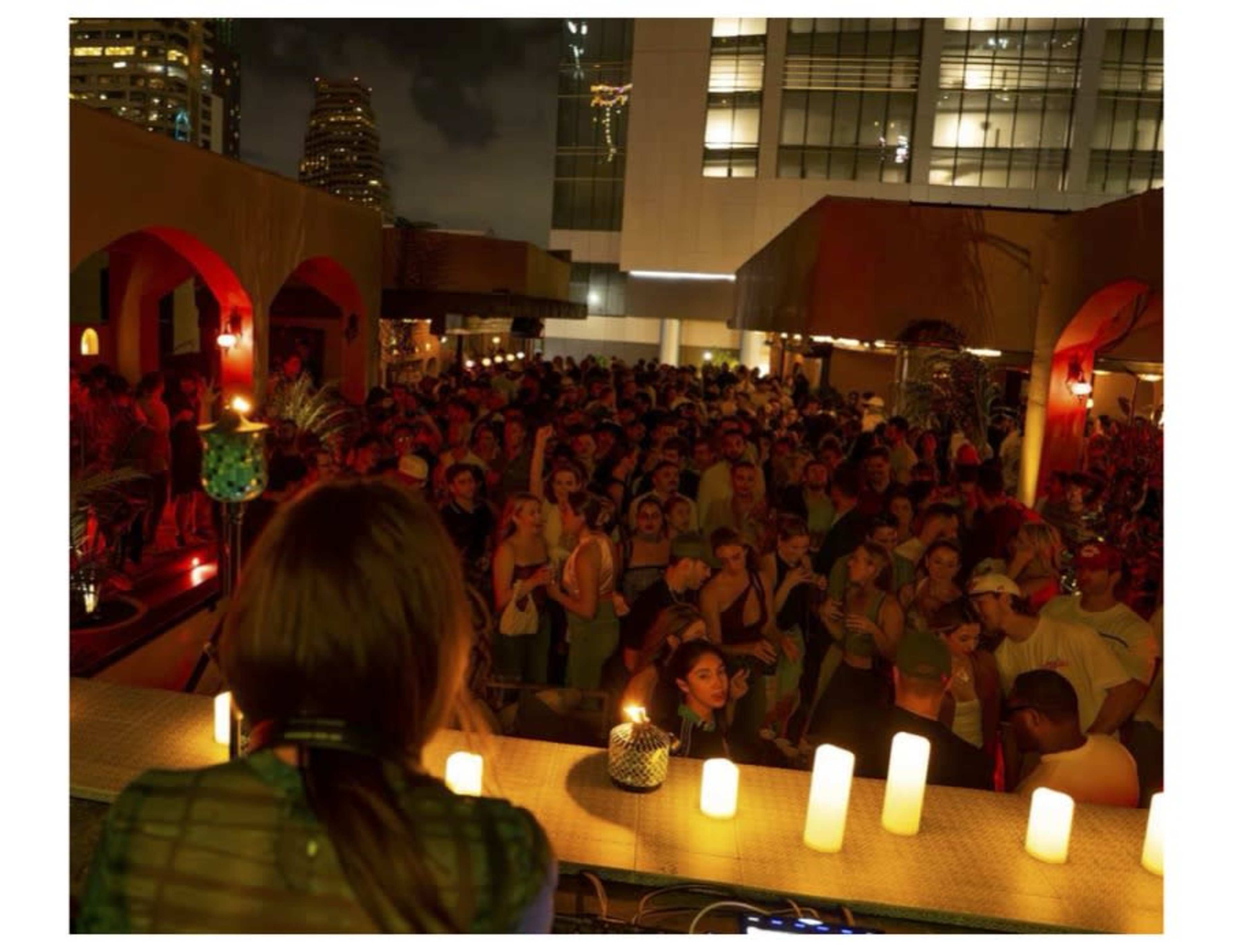 A crowded rooftop bar features a DJ with candles and colorful lighting, while a large audience enjoys the evening atmosphere.
