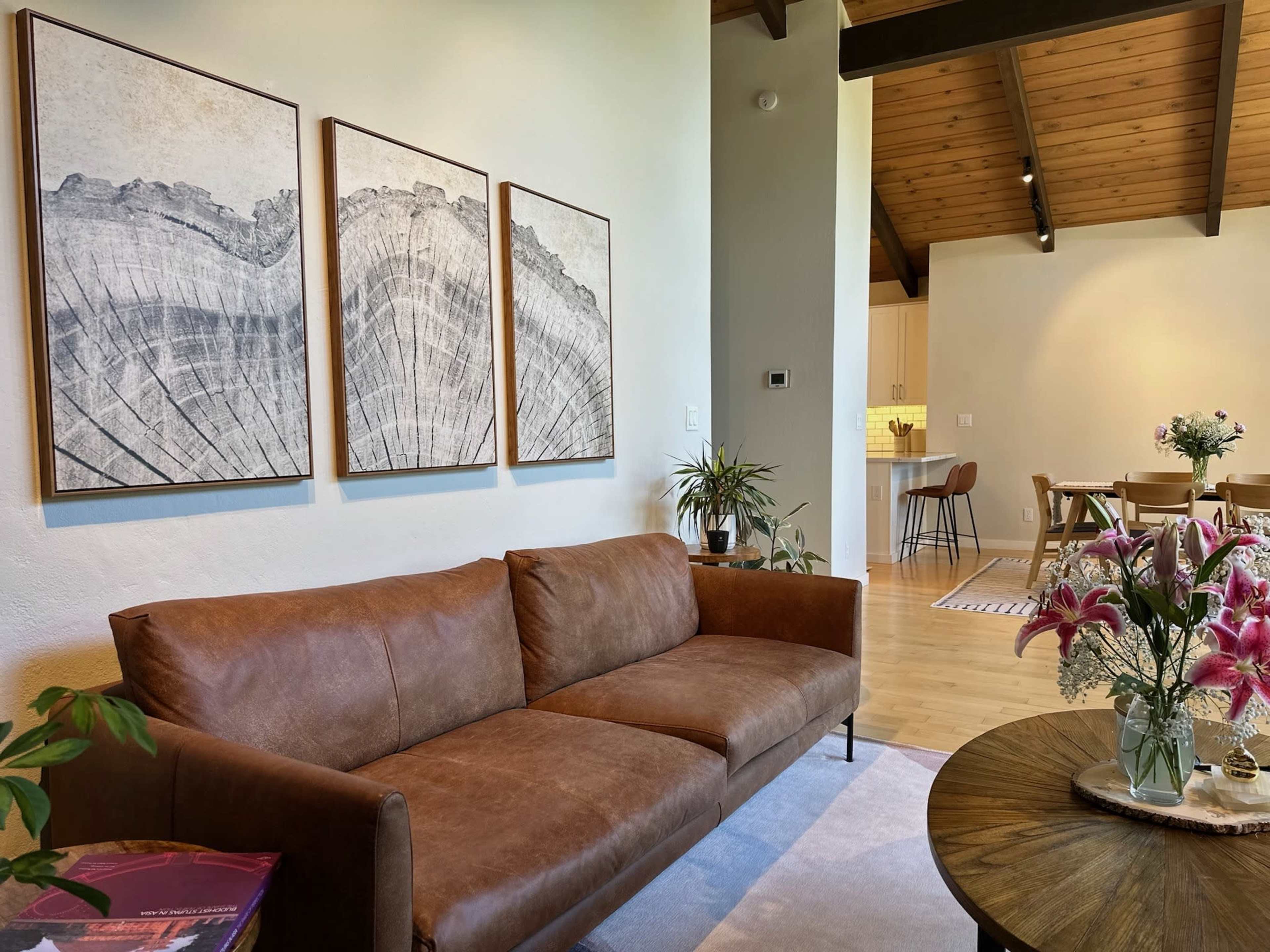 A living room featuring a brown leather sofa, three framed artworks on the wall, and a round coffee table with flowers and books.