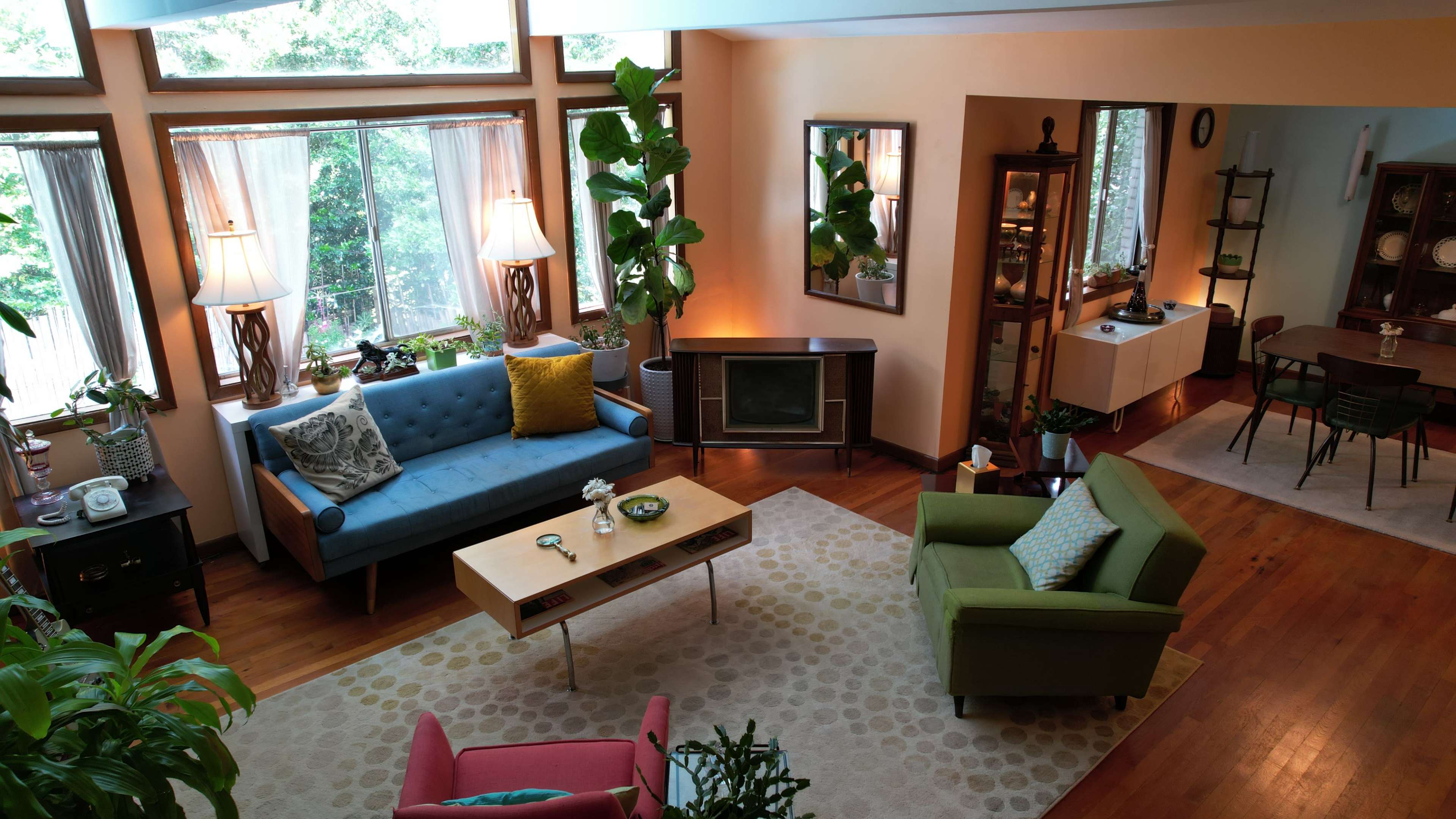A spacious living room with large windows, a blue sofa, a green armchair, a wooden coffee table, and various indoor plants.