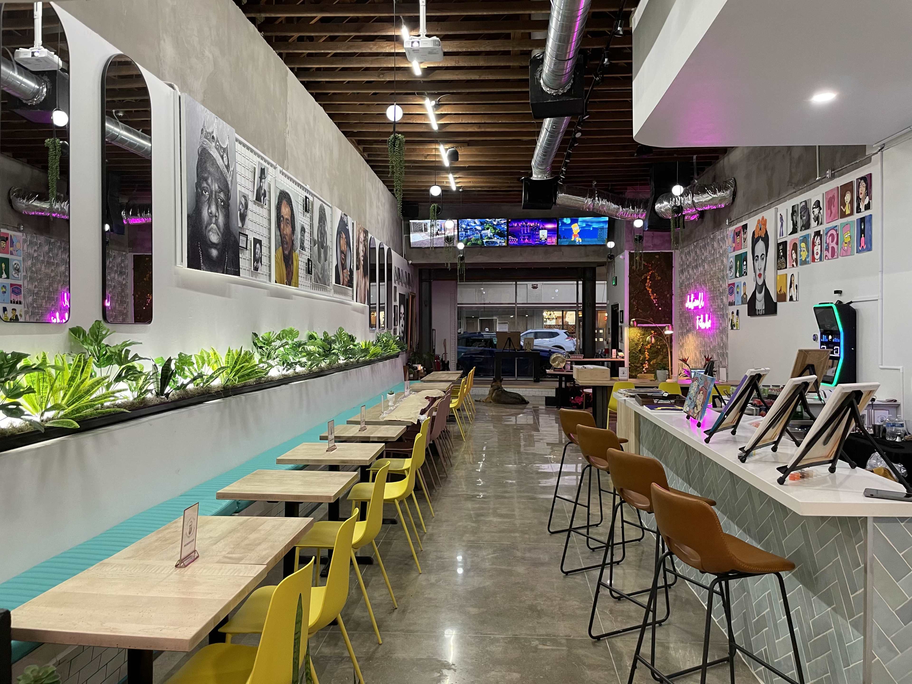 A modern restaurant interior with a long row of tables, colorful chairs, and walls adorned with photographs and greenery.