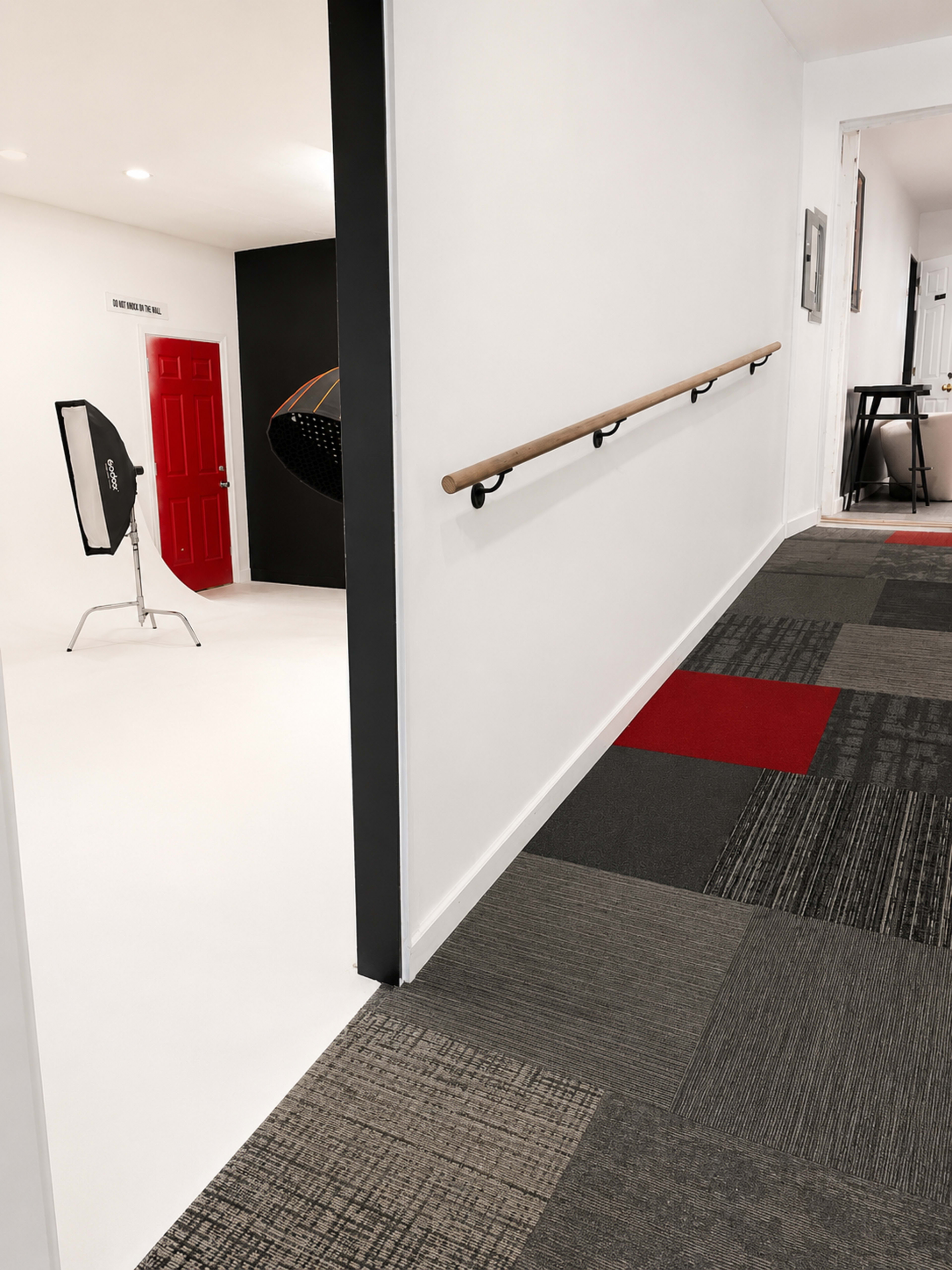 The image shows a corridor with a railing on the left side, leading to a brightly lit room with a red door and photography equipment in the background.