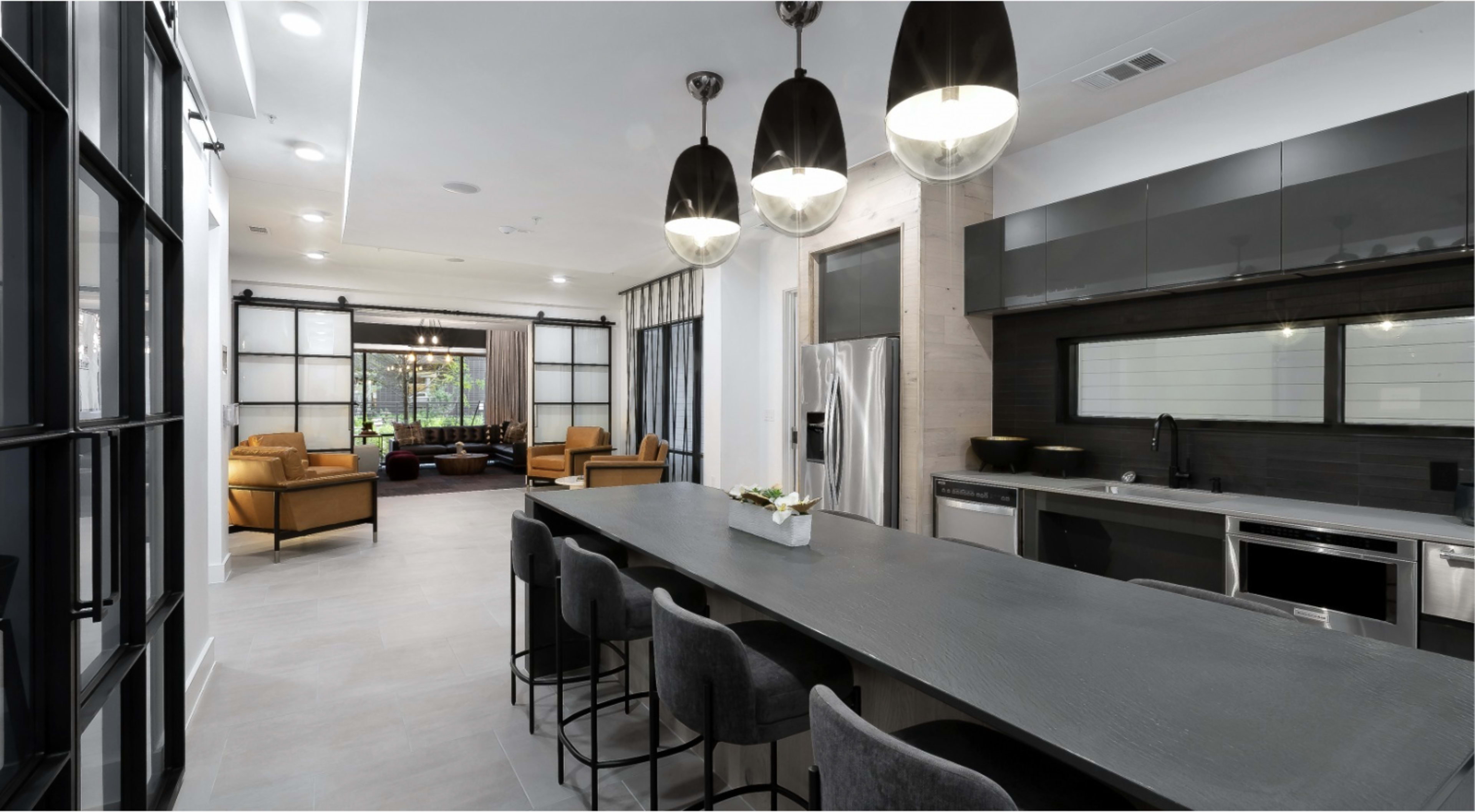 The image shows a modern kitchen with a long countertop and bar seating, adjacent to a living area featuring two armchairs and large windows overlooking a garden.