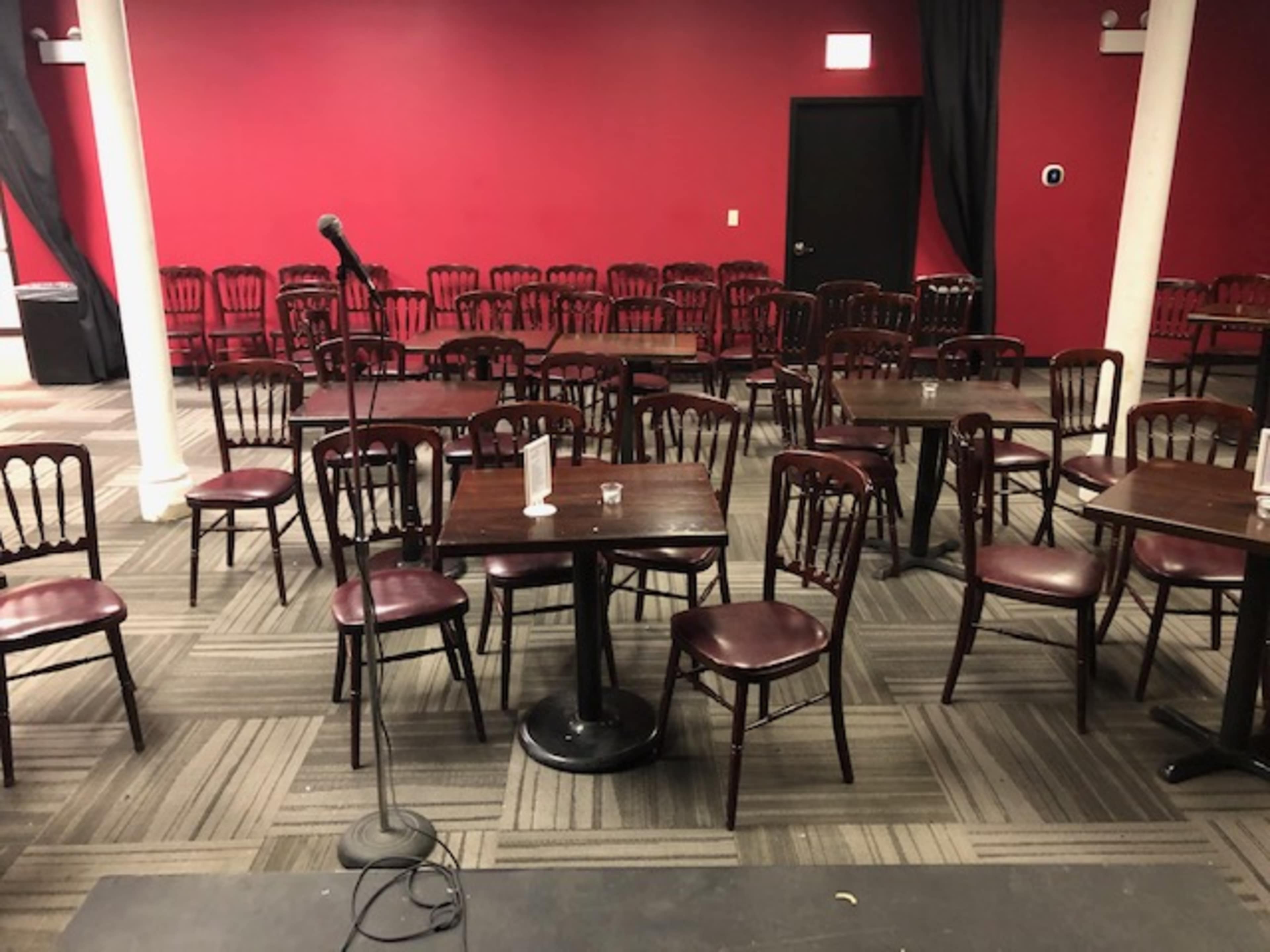 A microphone stands in front of a small stage facing a row of empty tables and chairs in a red-walled performance venue.