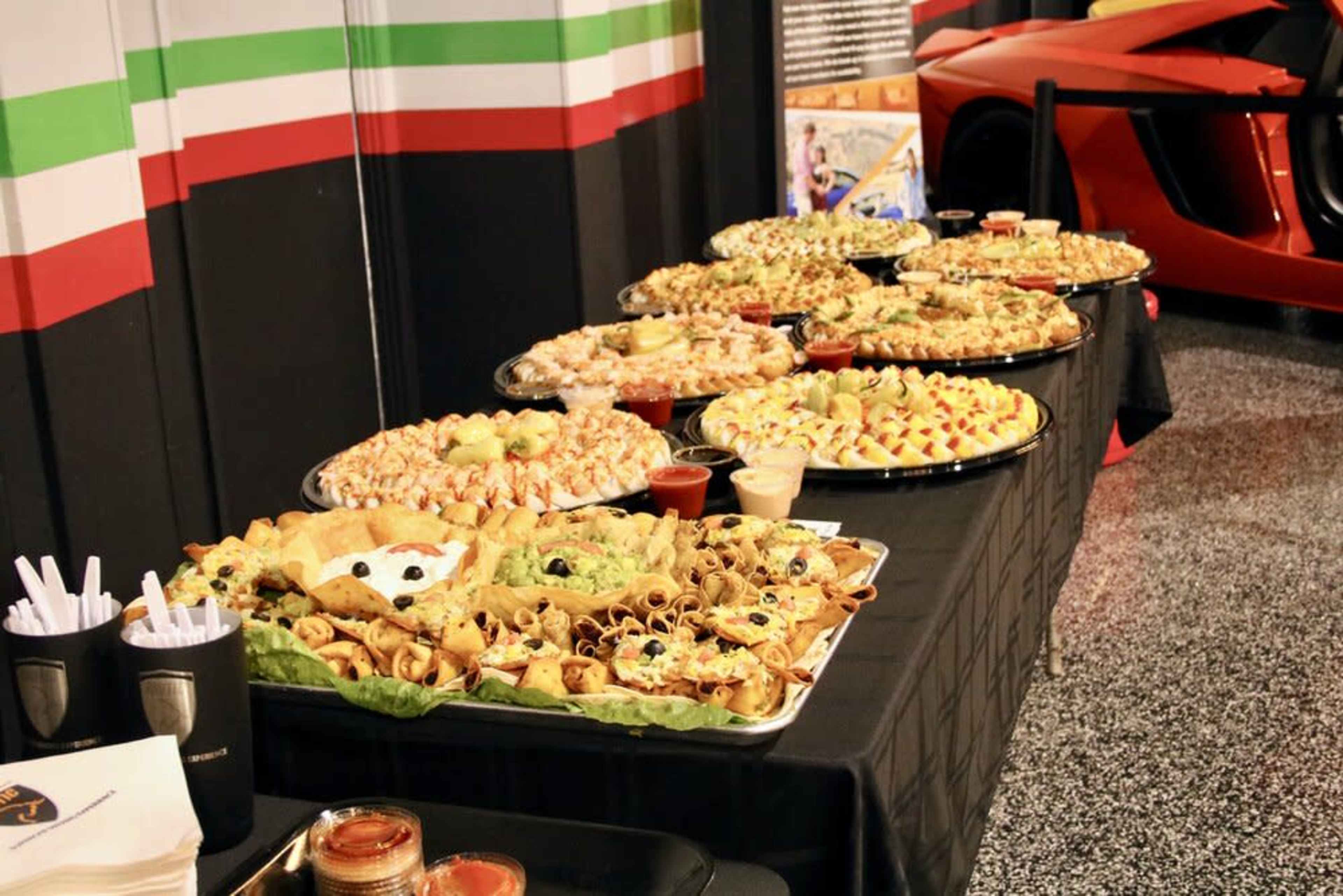 A long table covered with various platters of appetizers and snacks is set up next to a red sports car in a decorated venue.