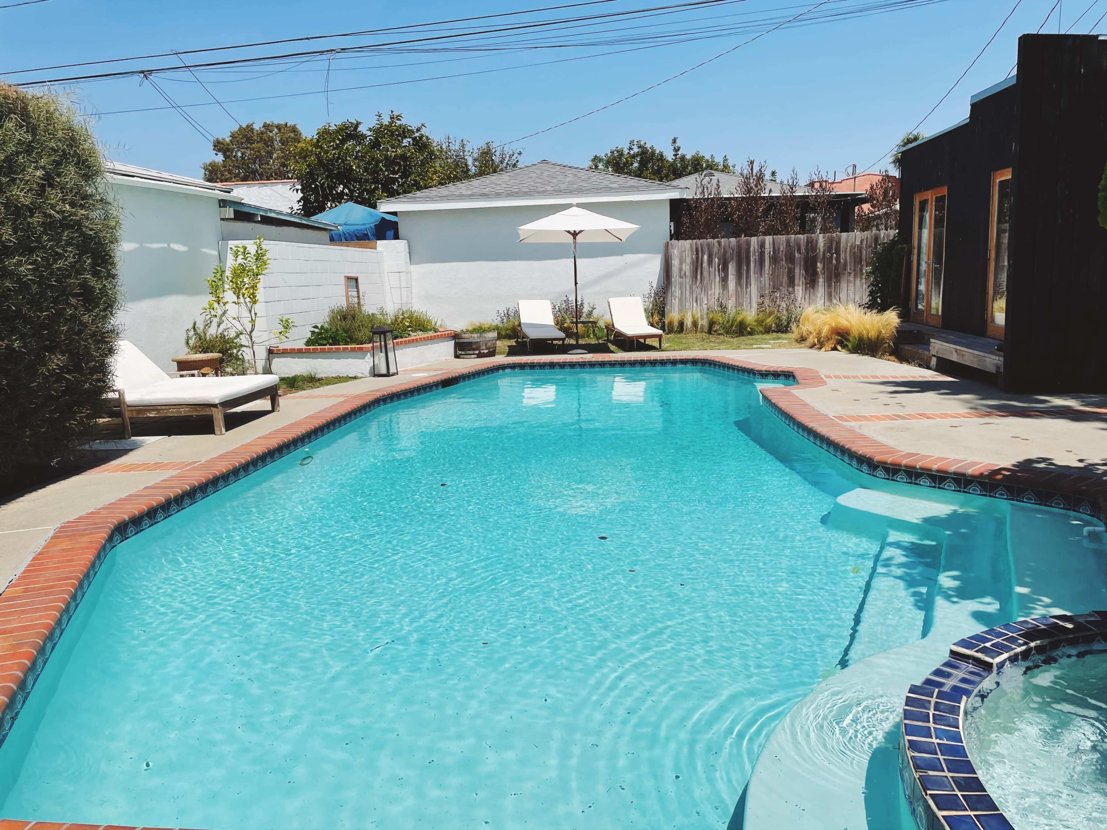 A clear blue swimming pool is surrounded by lounge chairs and a patio, with a shaded umbrella in the background and a wooden structure nearby.