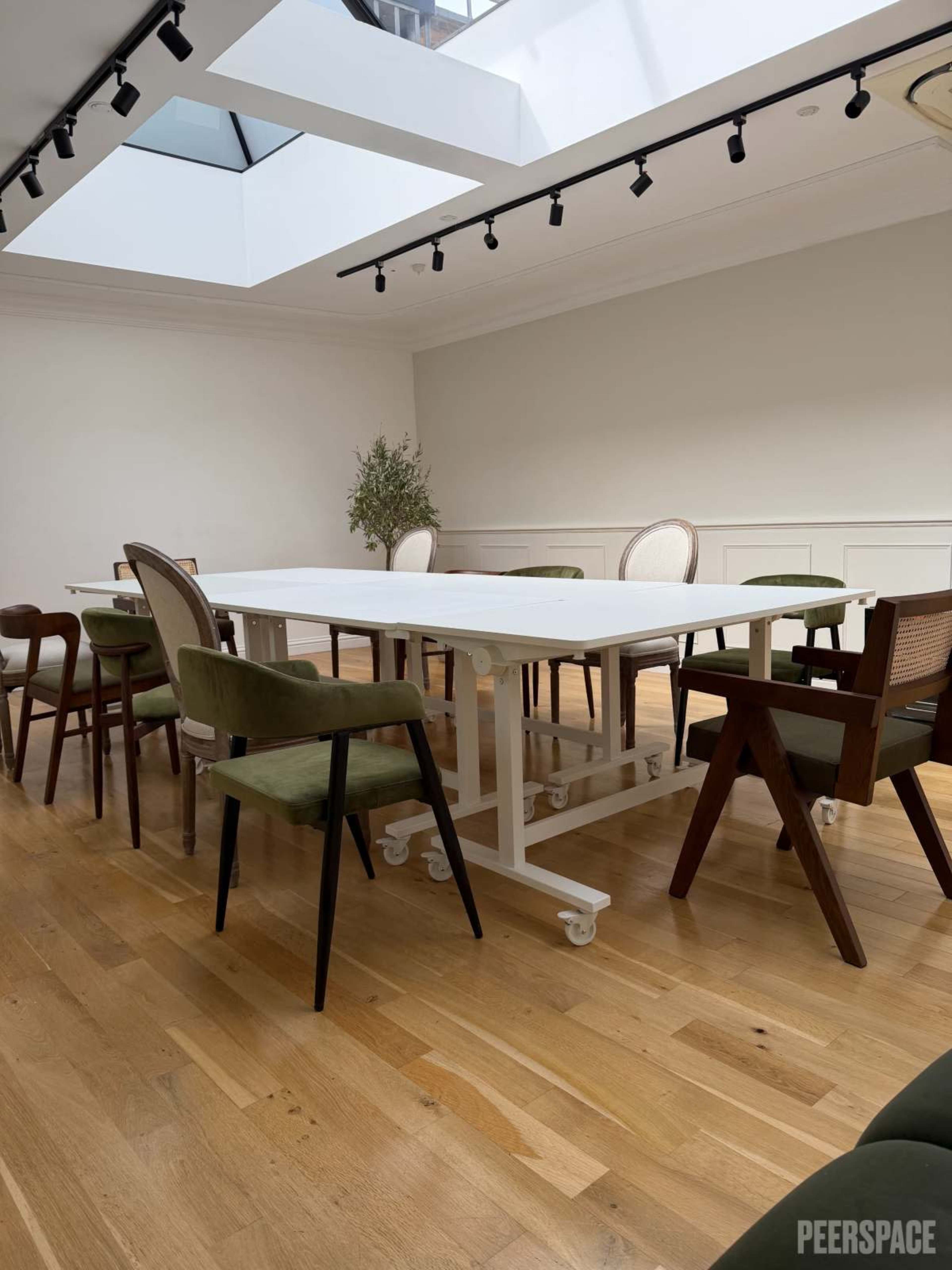 A large white table on wheels is surrounded by various chairs in a well-lit room with a skylight.