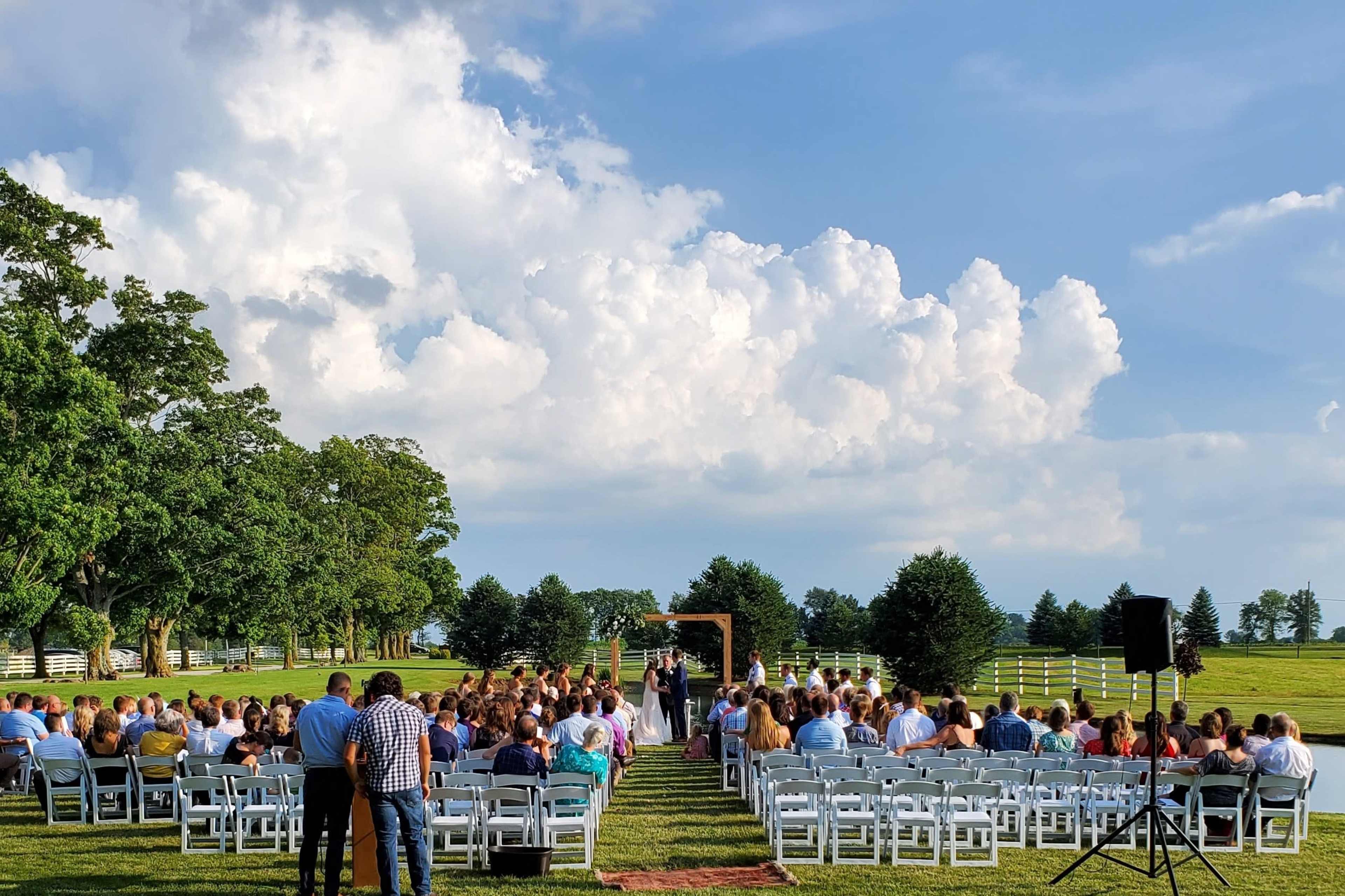 A wedding ceremony is taking place outdoors with white chairs arranged in rows facing an arch, under a bright sky filled with clouds.
