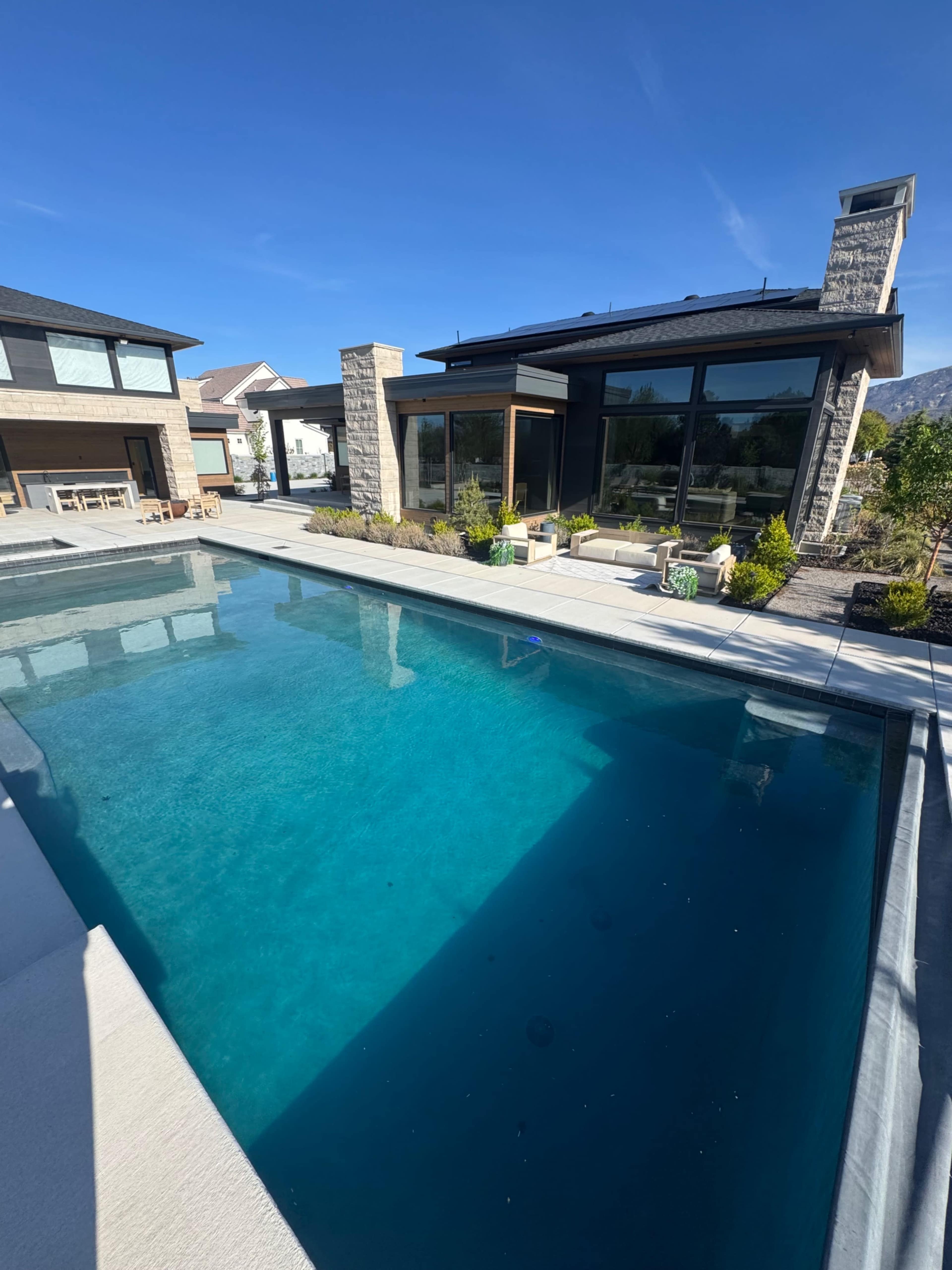 A clear blue swimming pool reflects the sky next to a modern house with large windows and a landscaped area.
