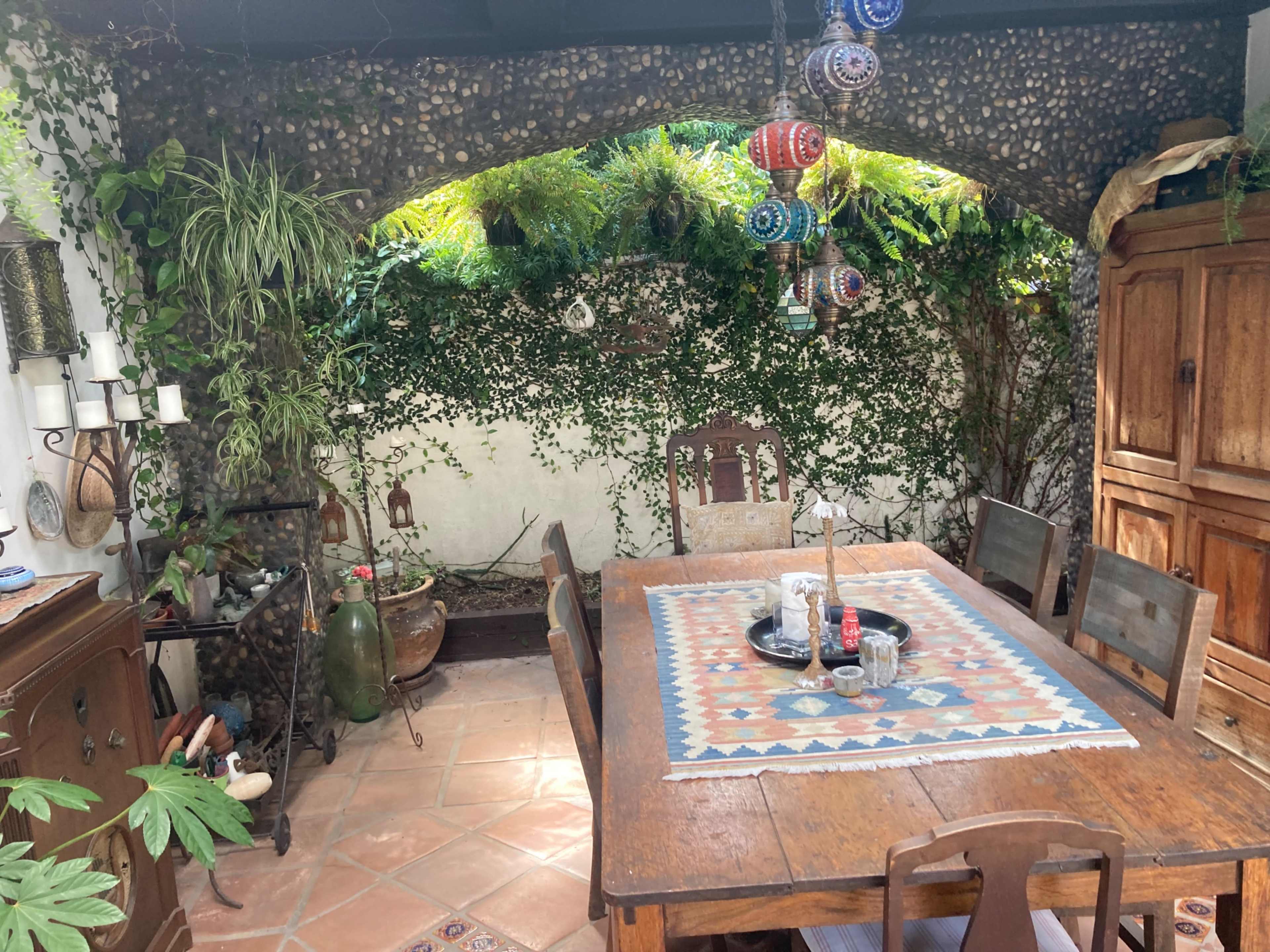 A wooden dining table is set in a shaded outdoor area surrounded by greenery, with colorful hanging lanterns above.