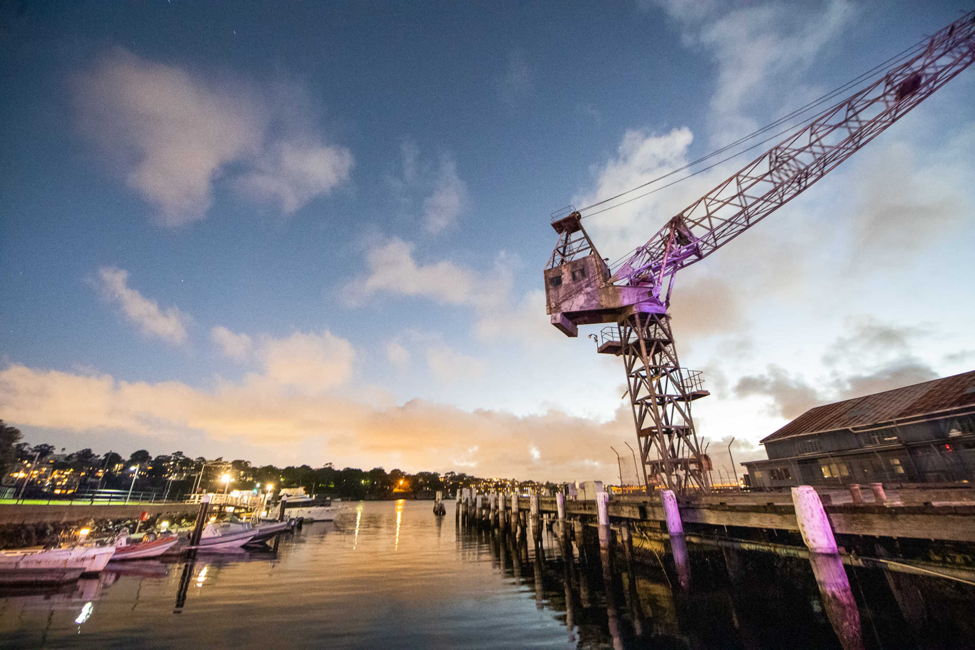 A large crane stands next to a calm body of water, with boats docked along weathered wooden pilings under a twilight sky.