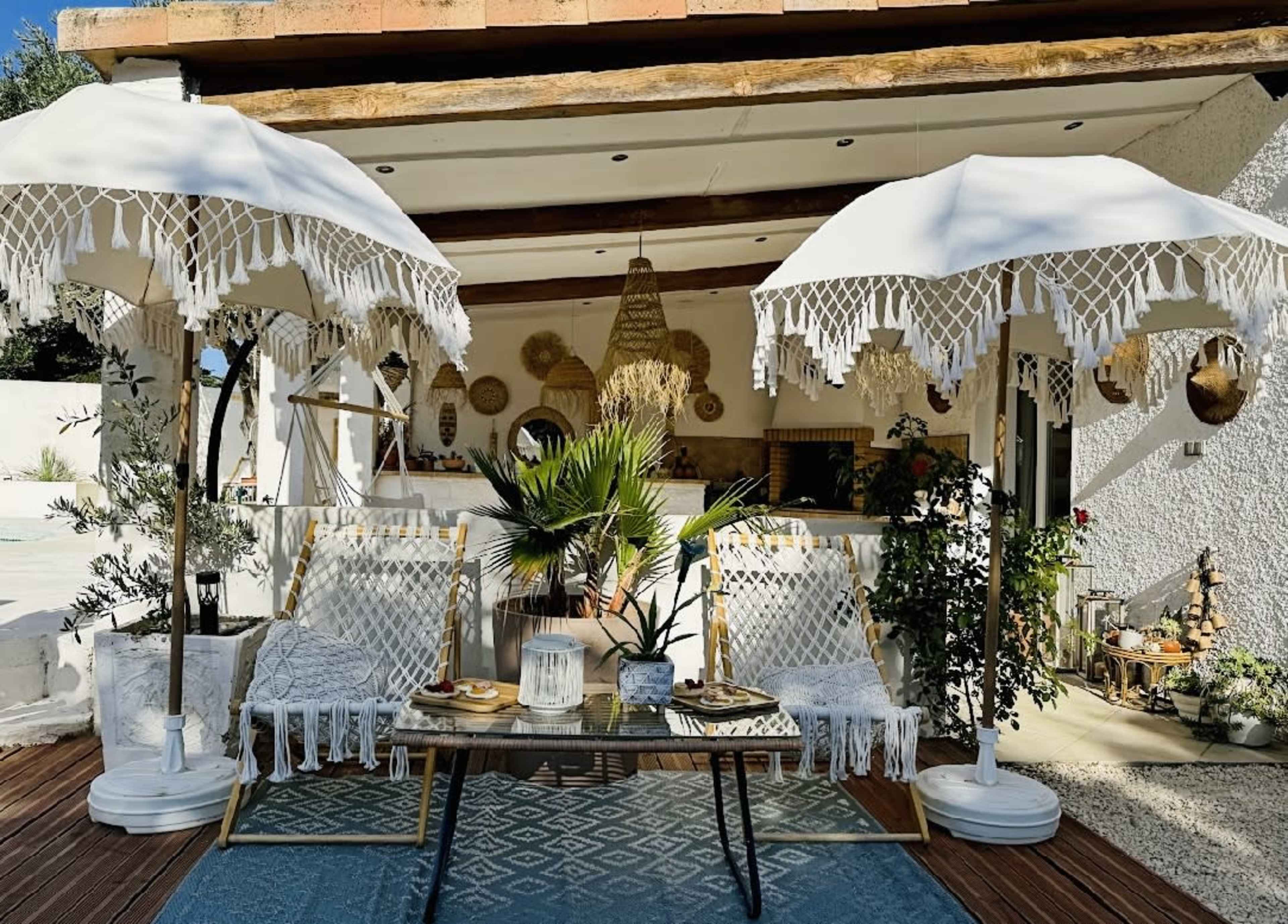 The image depicts a cozy outdoor seating area adorned with two white parasols, a low table, and several decorative plants in a sunlit setting.