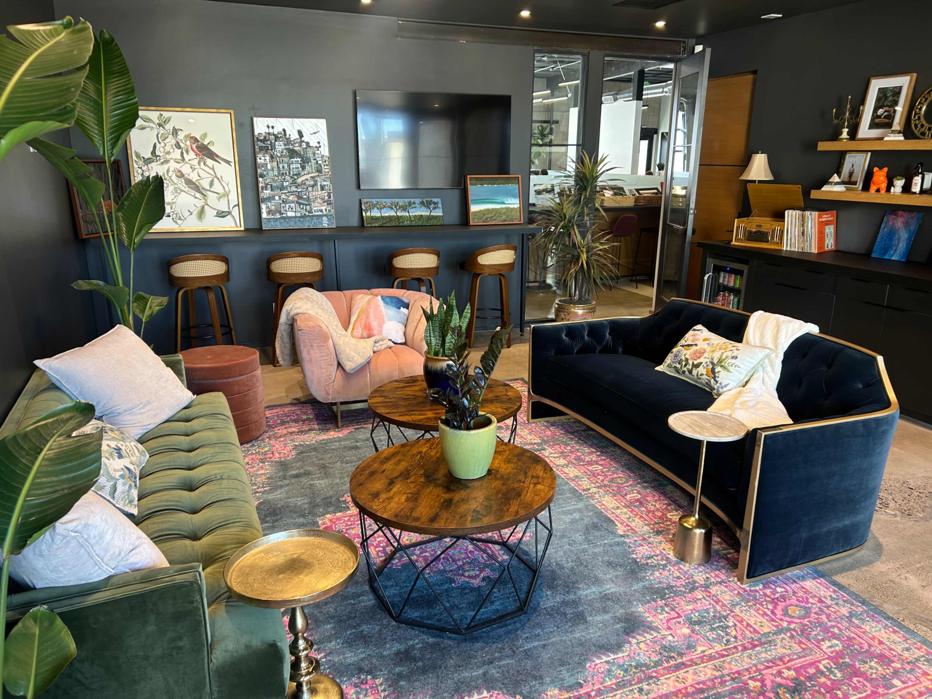 The image shows a modern lounge area with colorful sofas, circular coffee tables, and a decorative rug, accompanied by indoor plants and wall art.