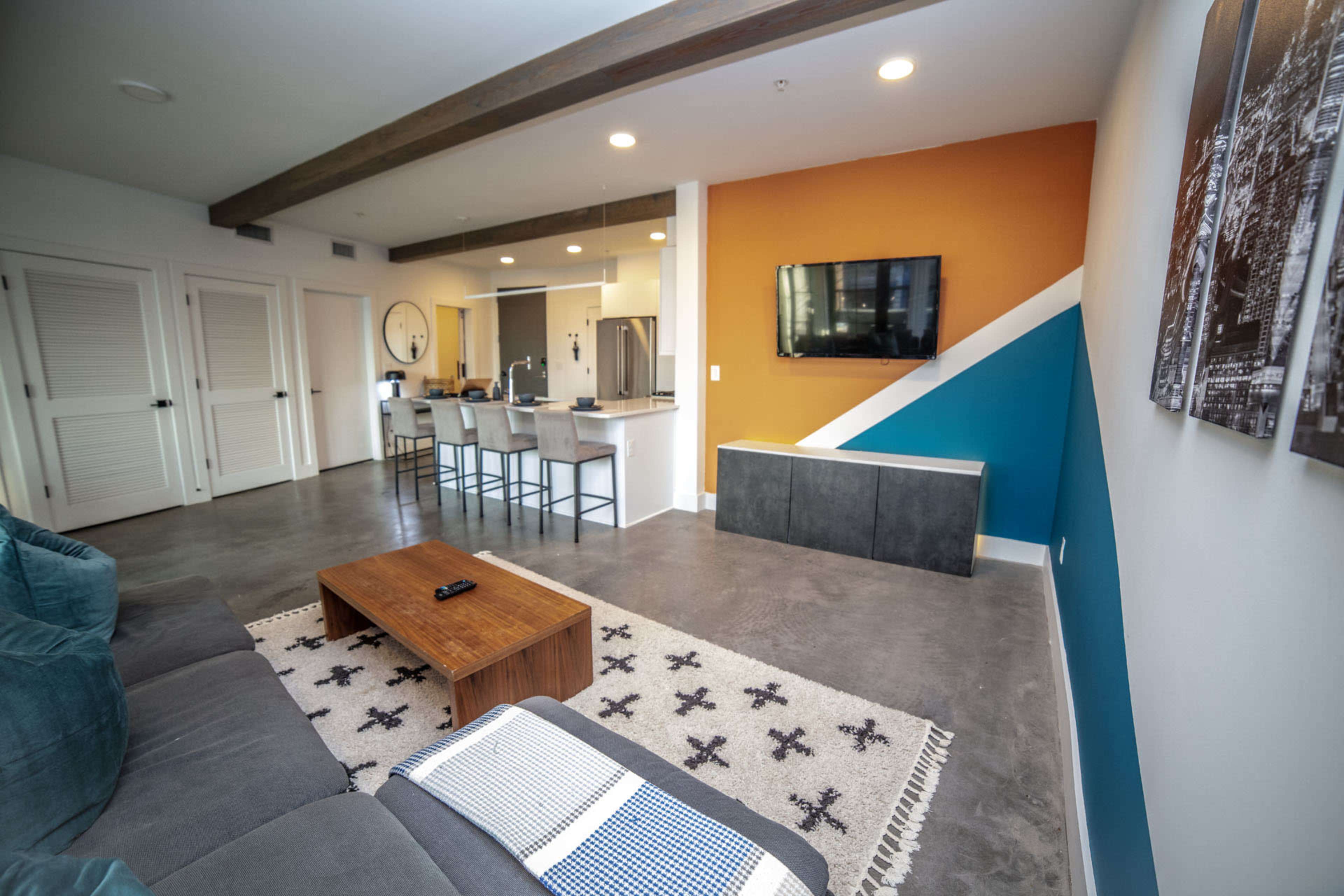 The image shows a modern living room with a gray sofa, a wooden coffee table, and a flat-screen TV mounted on a colorful wall.