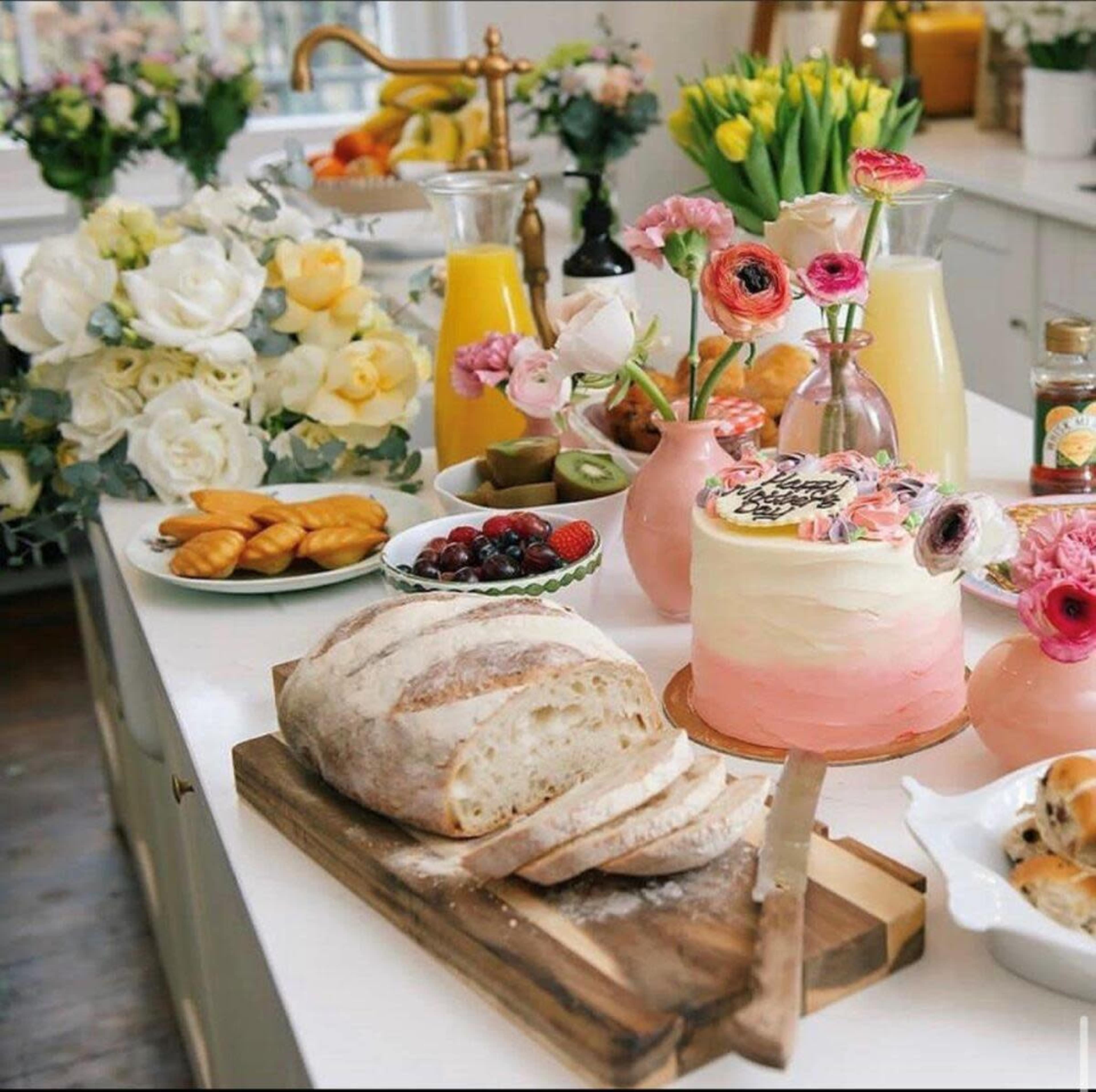 A variety of food items, including bread, cake, fruit, and drinks, are displayed on a kitchen counter surrounded by flowers.