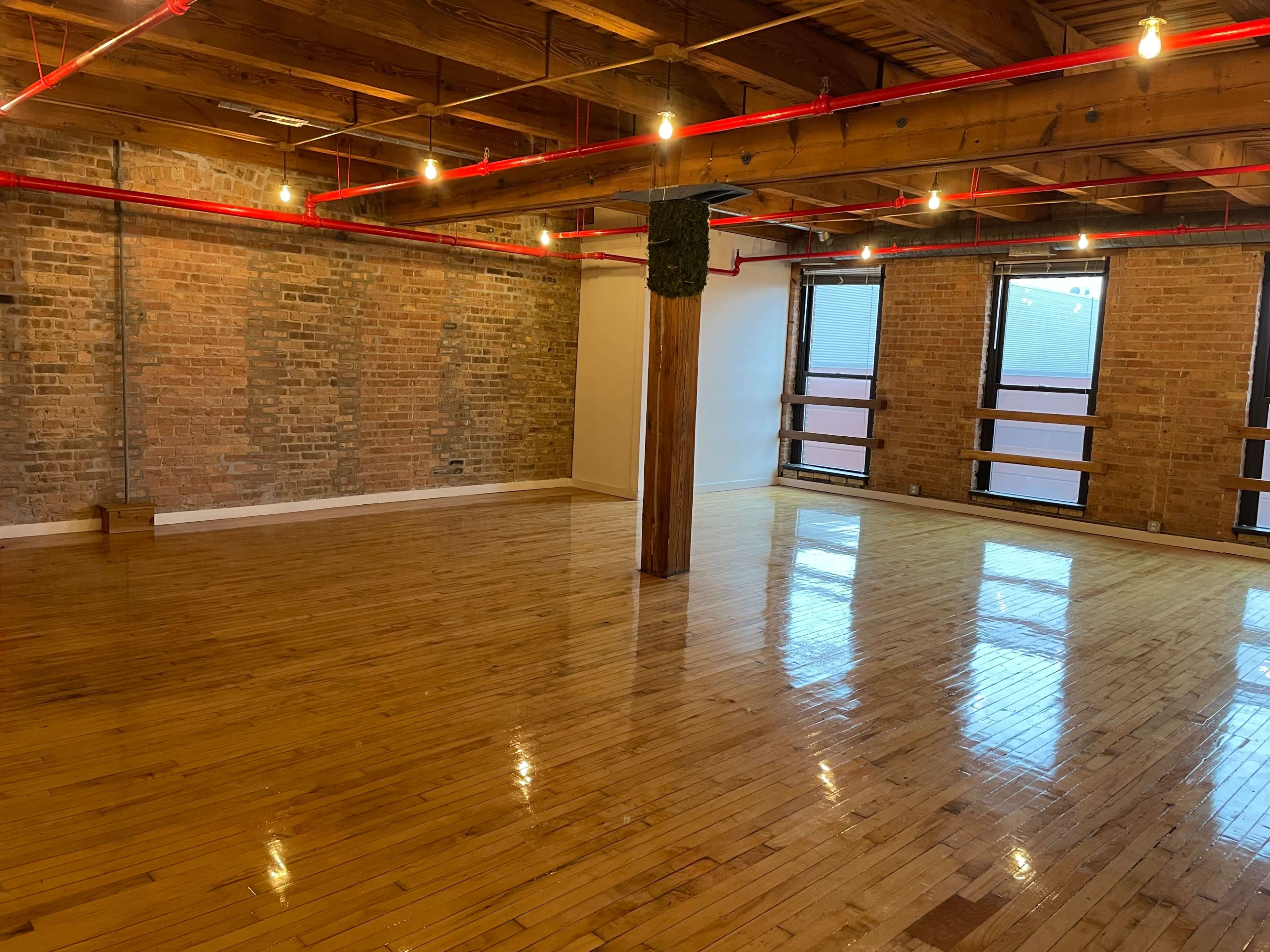 The image shows a spacious room with polished wooden floors, exposed brick walls, and red pipes running across the ceiling.