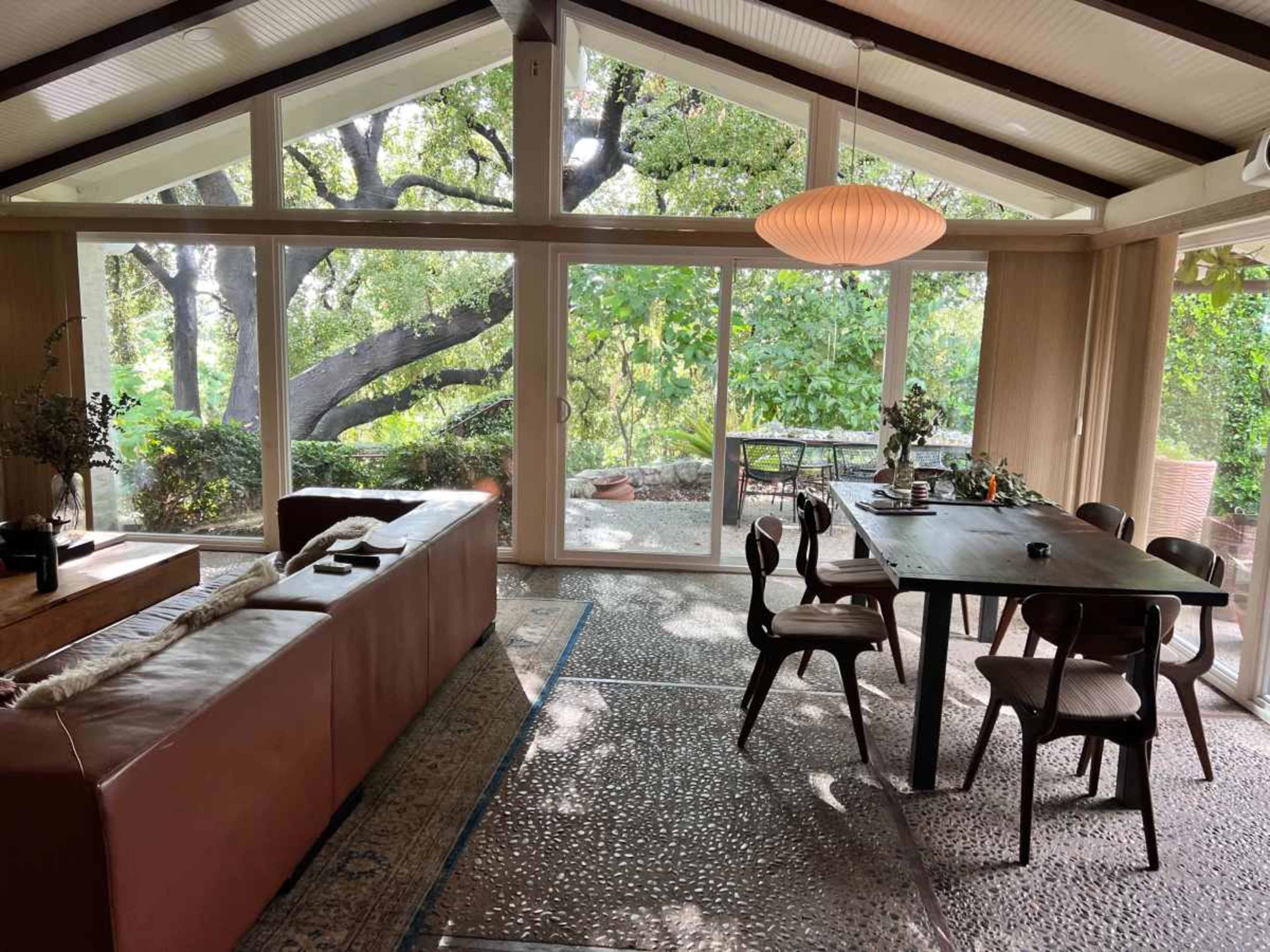 A spacious dining area with a wooden table and chairs opens up to a view of trees through large glass sliding doors.