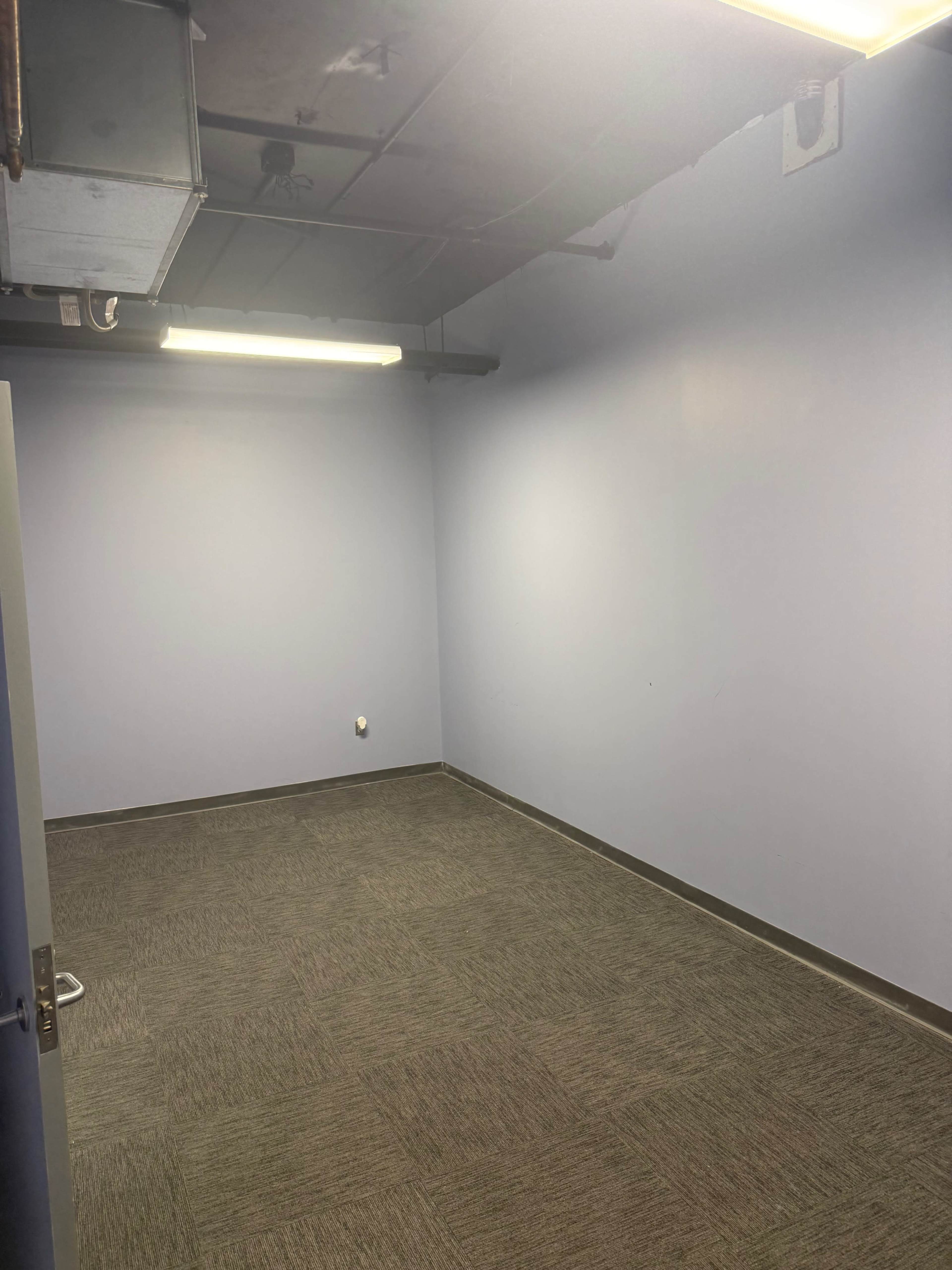 The image shows an empty room with light blue walls, a carpeted floor, and fluorescent lighting.