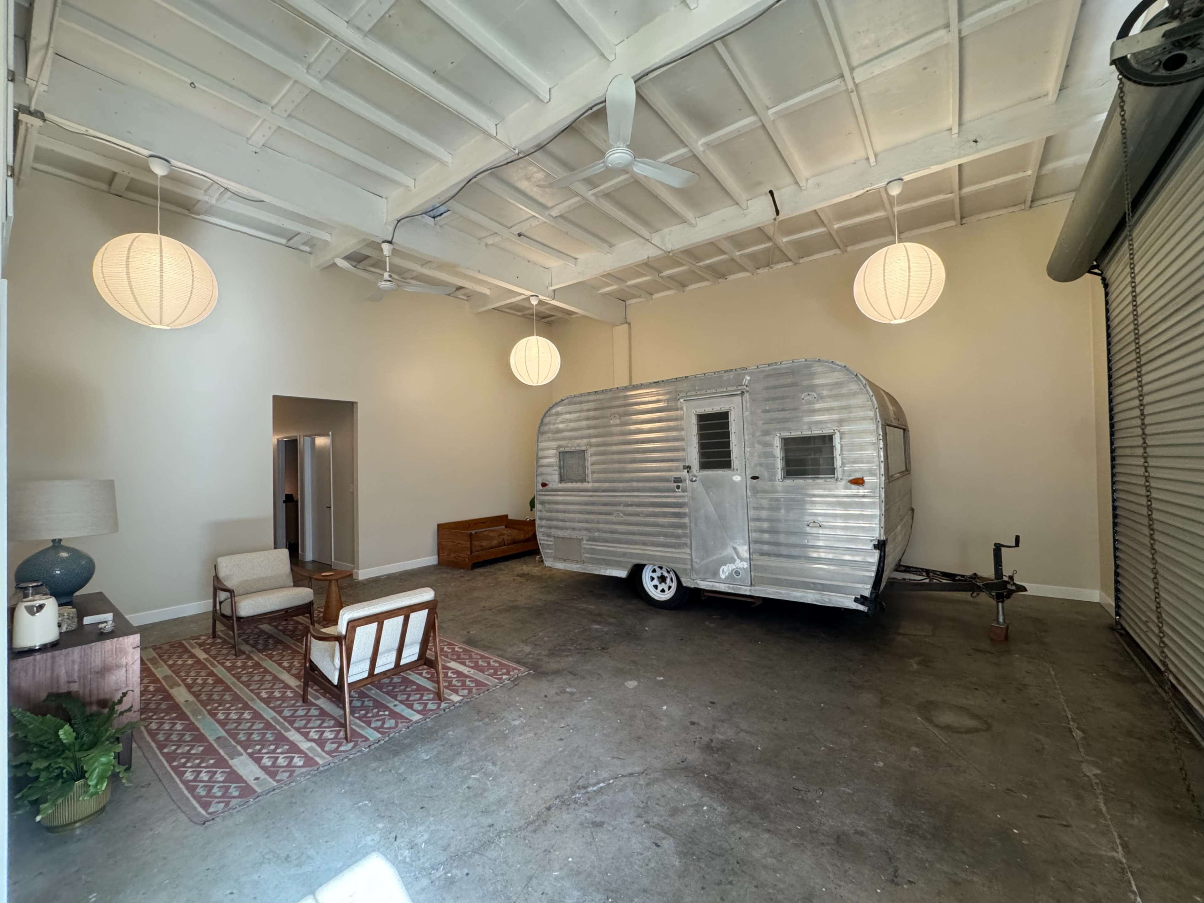 A silver vintage trailer is parked inside a spacious, brightly lit room with a simple seating area and decorative pendant lamps.