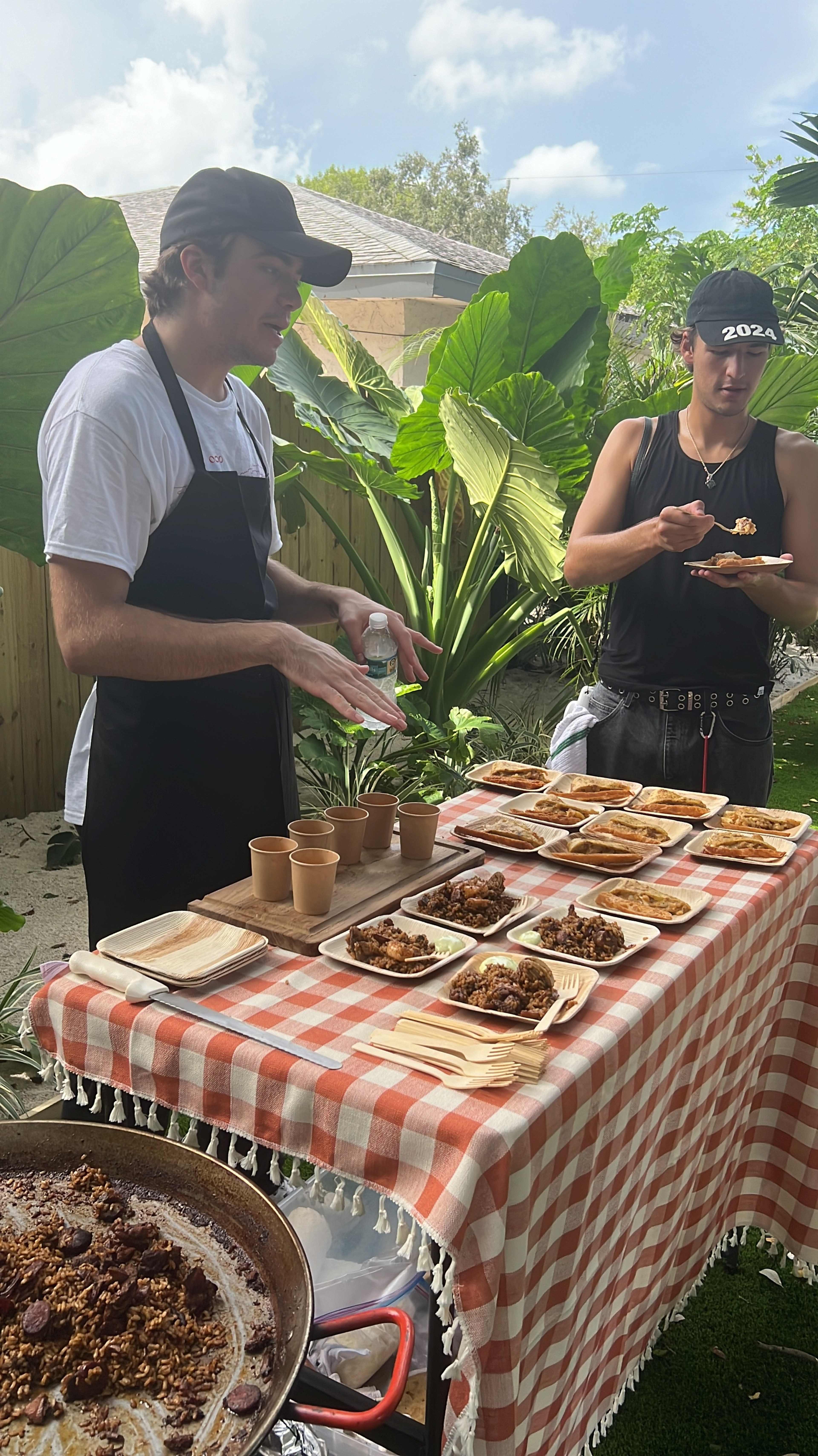 Two men stand behind a table covered with a checkered cloth, displaying plates of food alongside compostable cups in a lush garden setting.