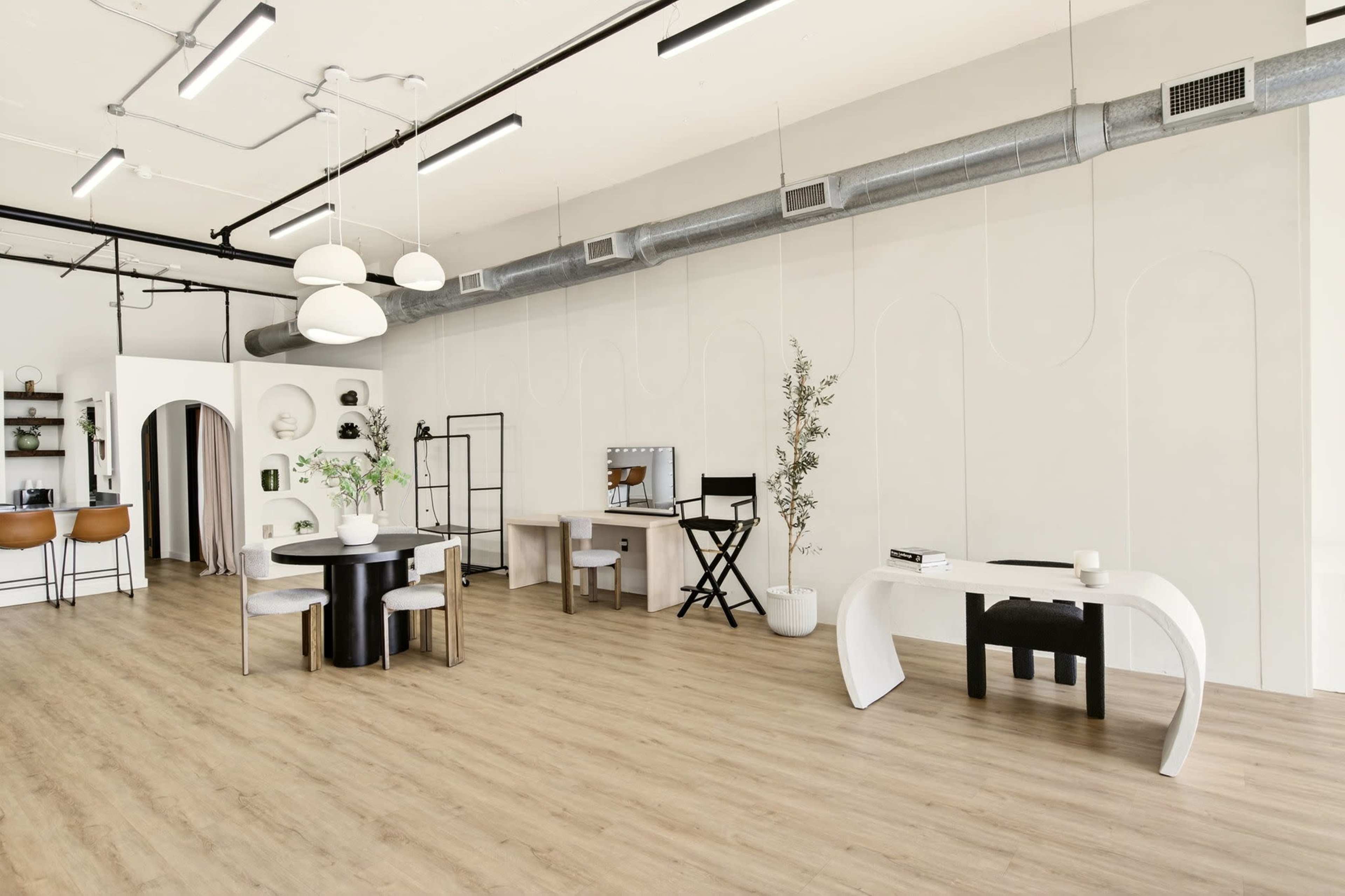 The image shows a modern, minimalist interior space featuring various workstations, with a mix of furniture styles and arrangements.