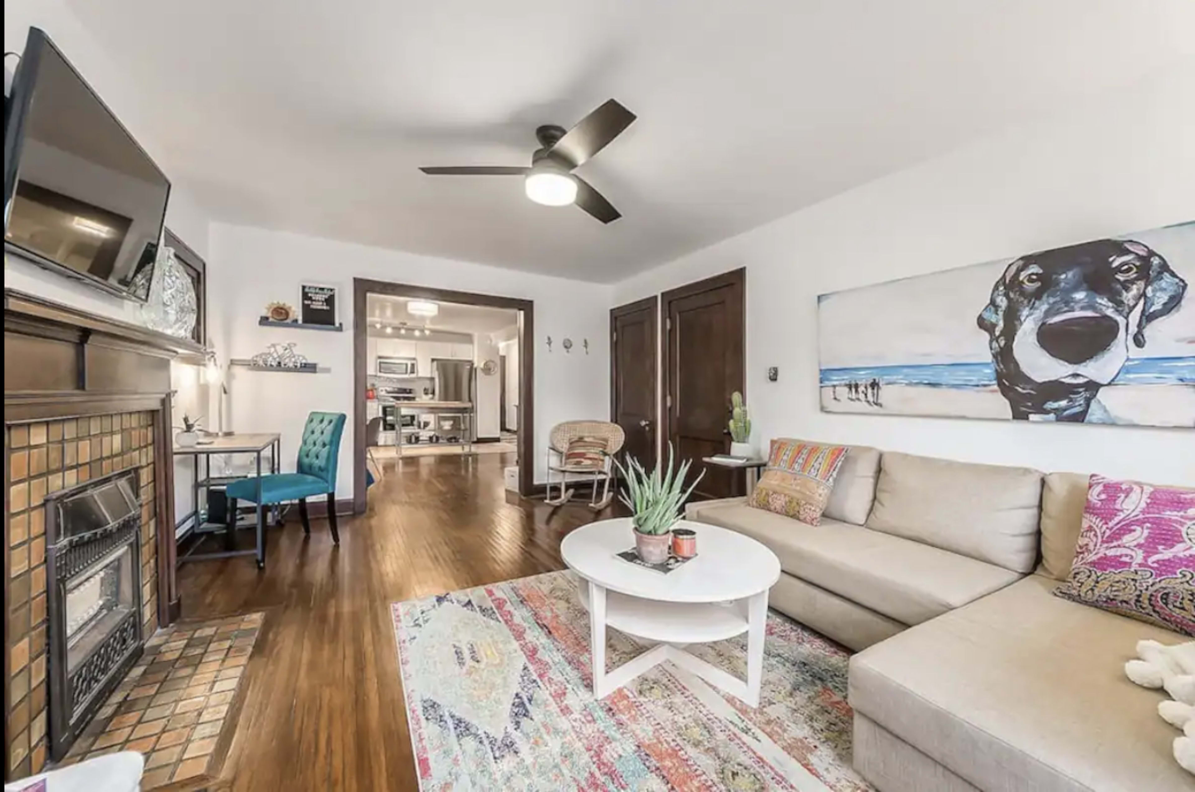 A living room with a beige sectional sofa, a round coffee table, a decorative rug, a wall-mounted television, and a large painting of a dog.