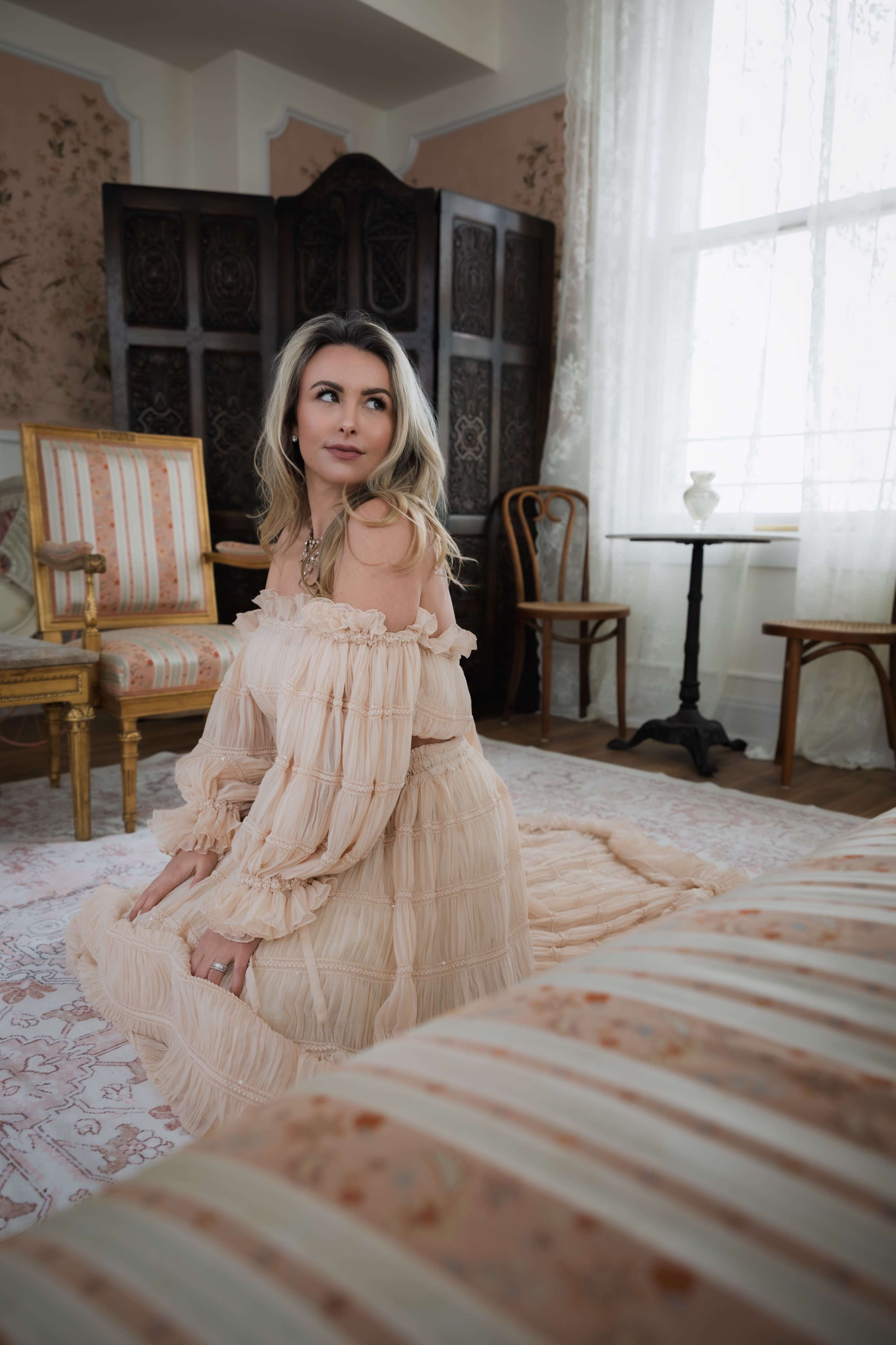 A woman in a light-colored off-the-shoulder dress is sitting on a plush rug in a vintage-style room with ornate furniture and large windows.