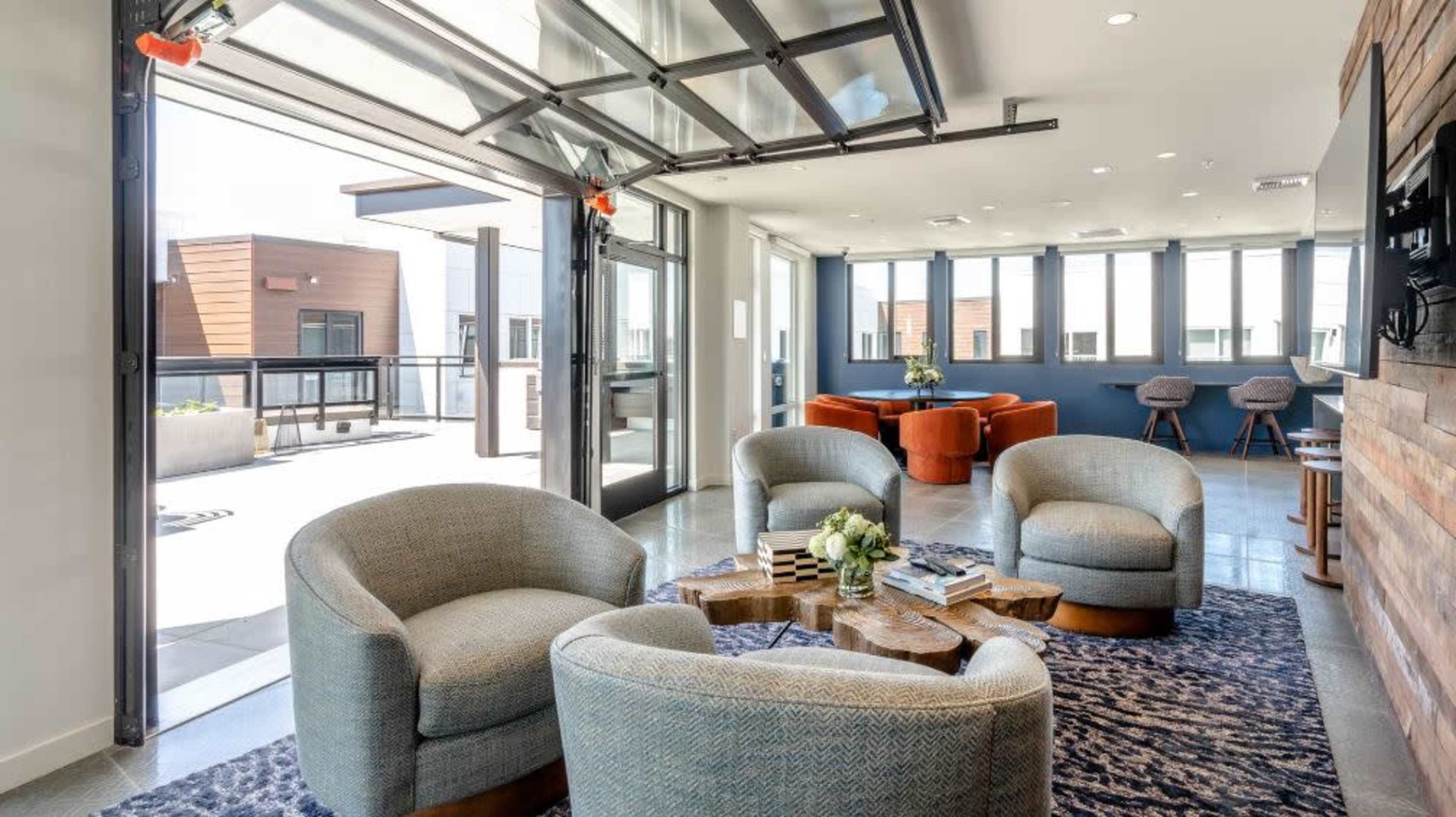 A modern lounge area with four curved chairs around a wooden coffee table, large windows, and a glass garage door that opens to an outdoor space.