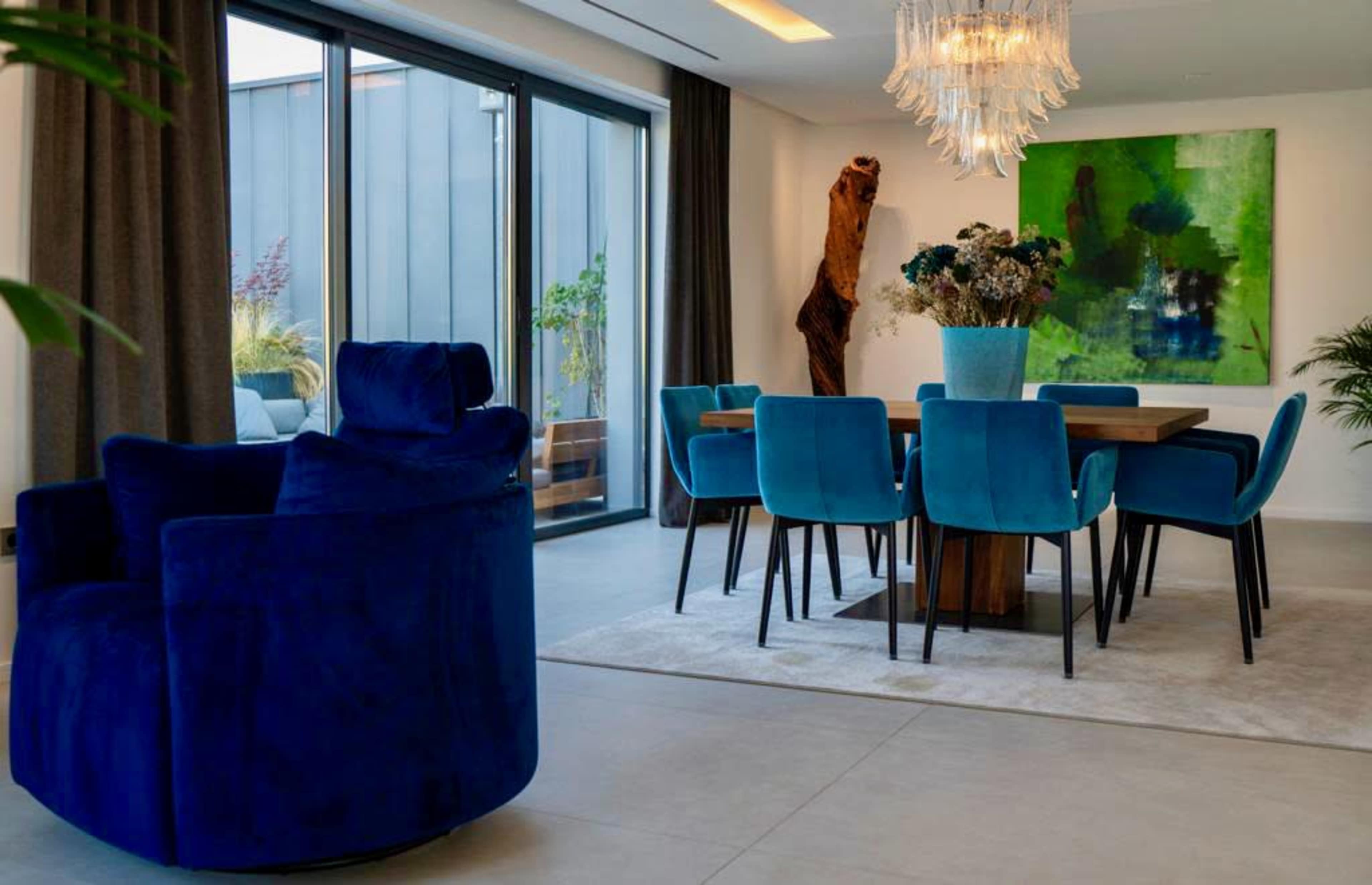 A modern dining area features a large wooden table surrounded by blue upholstered chairs and a decorative vase on the table, highlighted by a chandelier and an abstract painting on the wall.