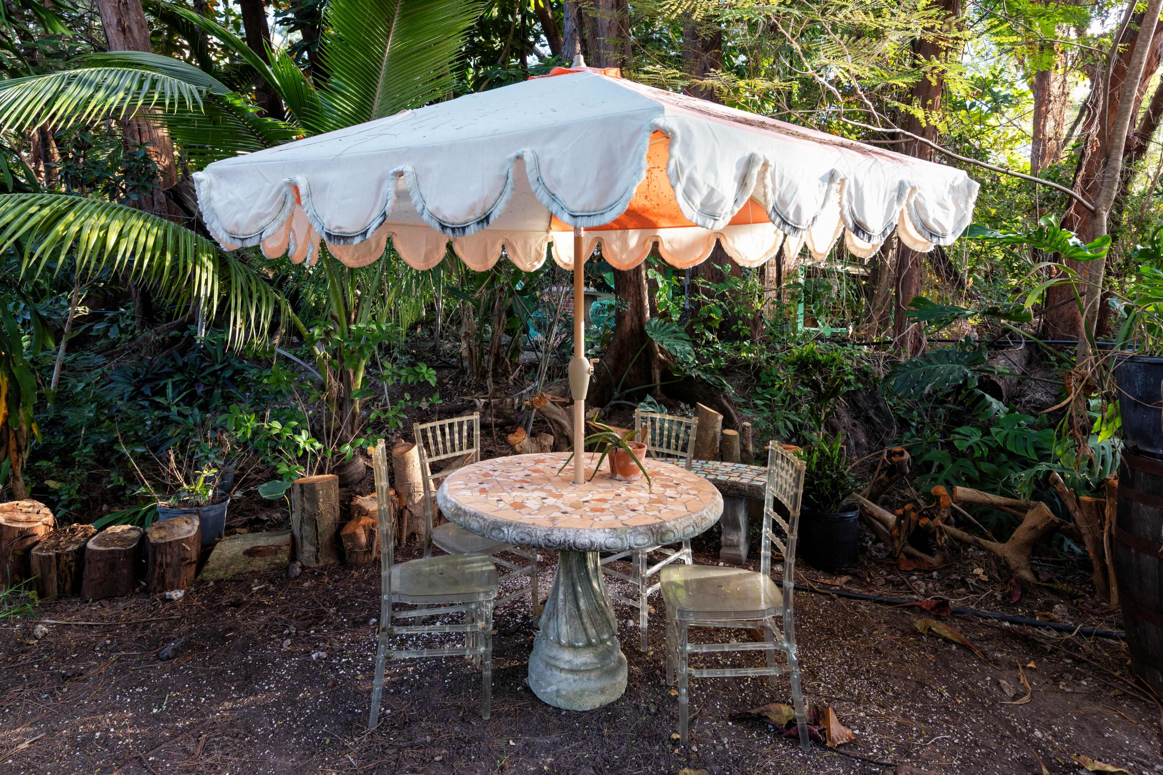 A table with a decorative umbrella and four chairs is set in a lush, green outdoor area surrounded by trees.