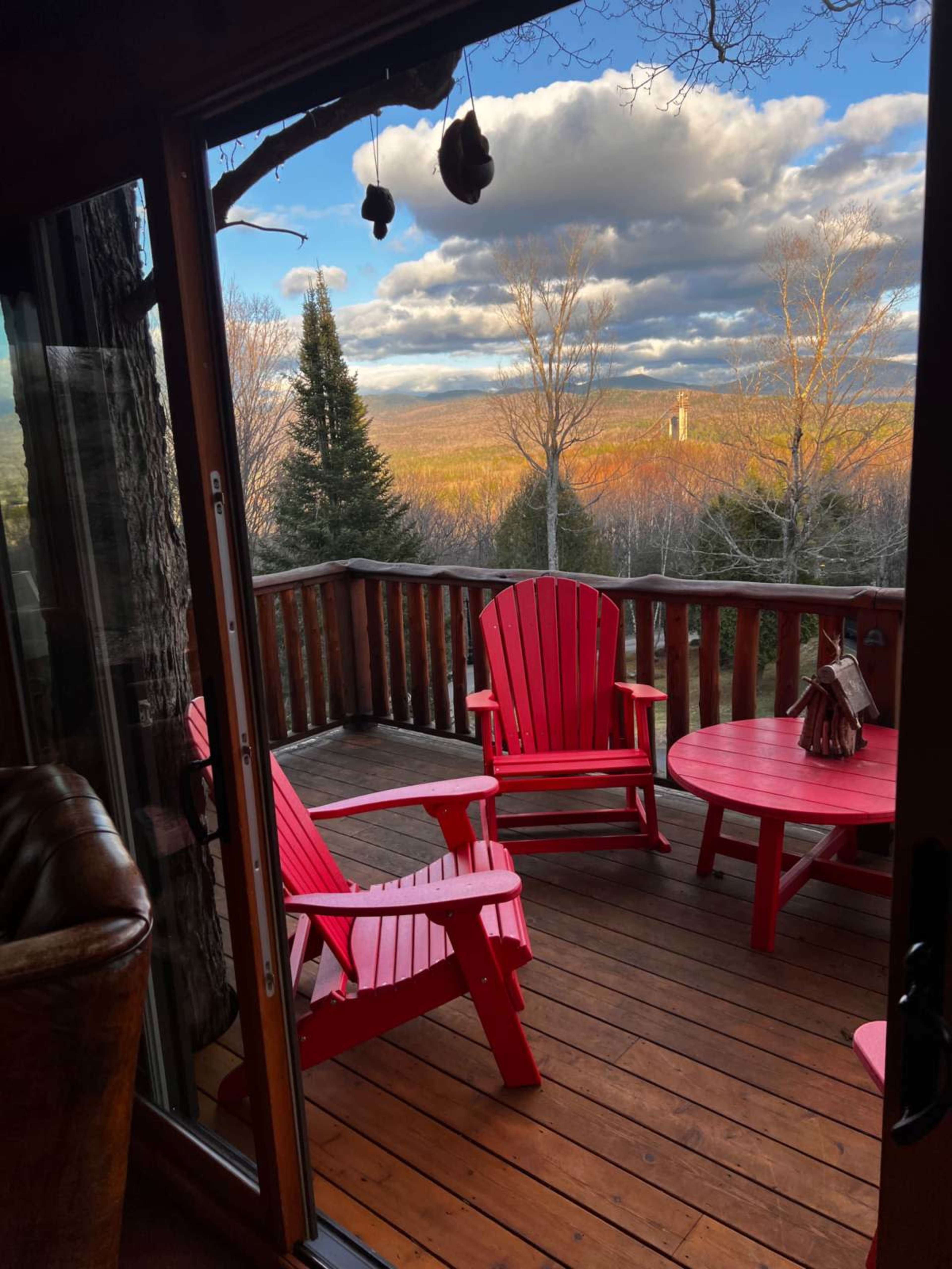 A wooden deck features two red Adirondack chairs and a round table, overlooking a valley with trees and distant mountains under a cloudy sky.
