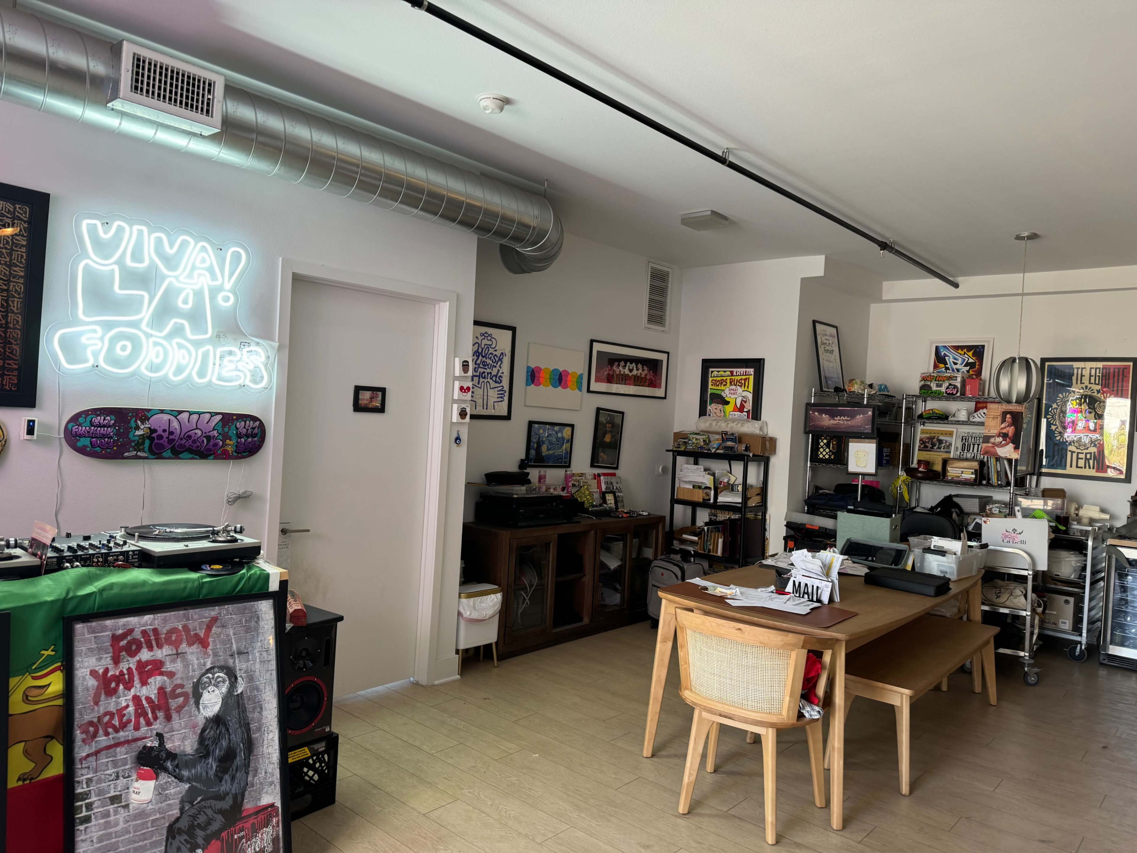 The image shows an eclectic room featuring a dining table, a variety of artwork on the walls, a record player setup, and a neon sign that reads "Viva! La! Foodies."