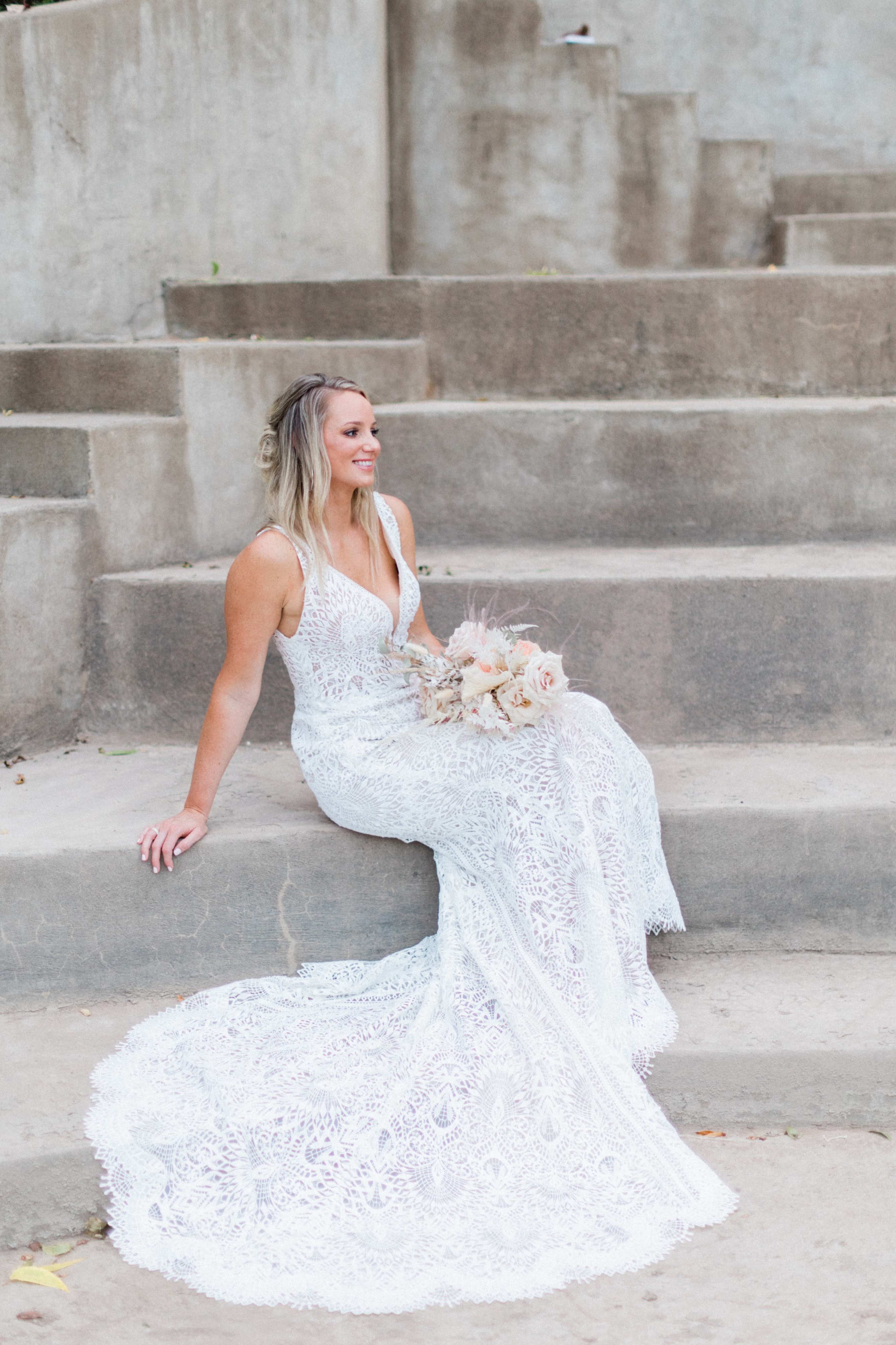 A woman in a lace wedding gown sits on a set of concrete steps, holding a bouquet of flowers.
