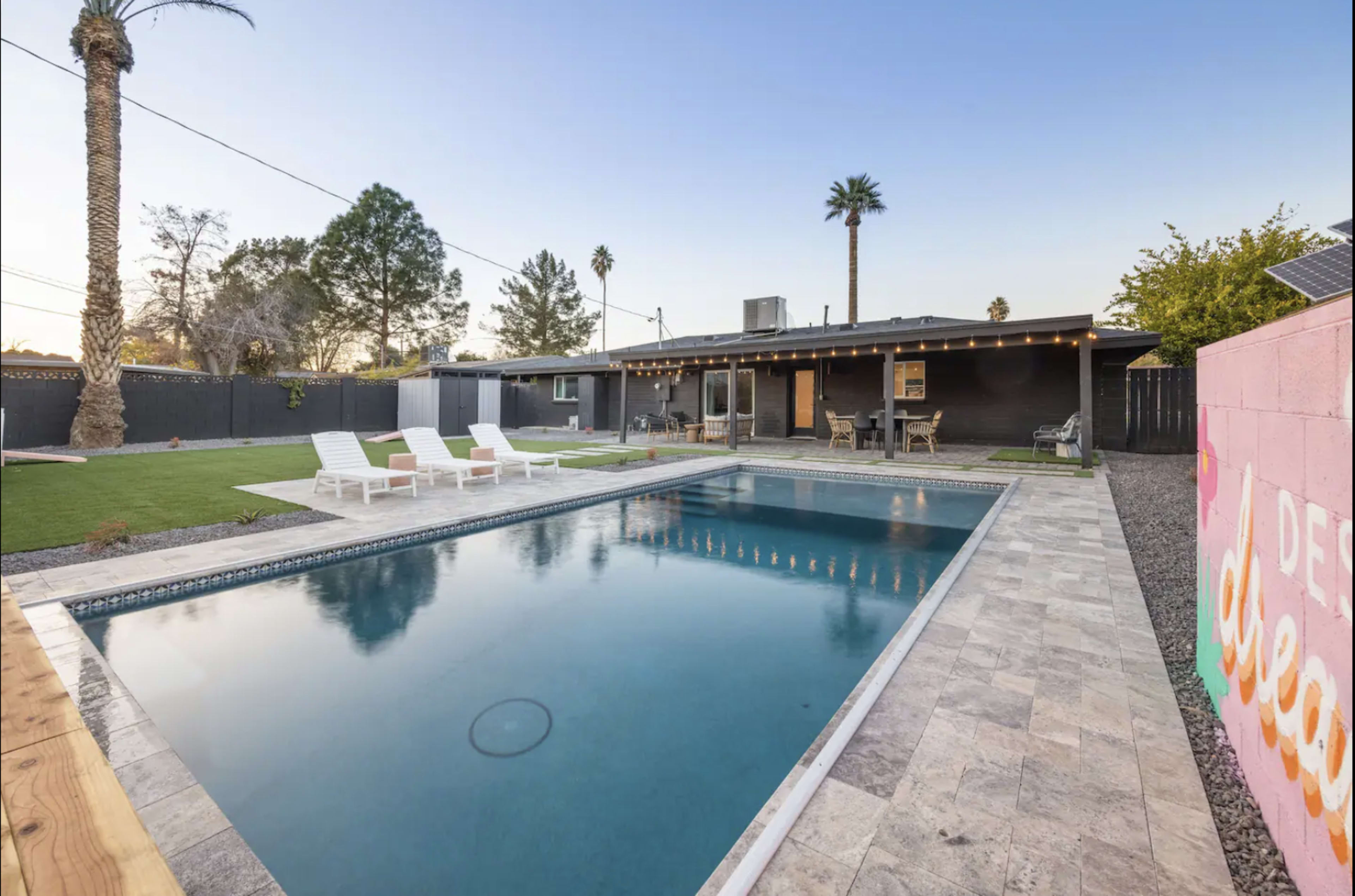 A backyard with a swimming pool, lawn chairs by a swimming pool, and a modern house in the background.