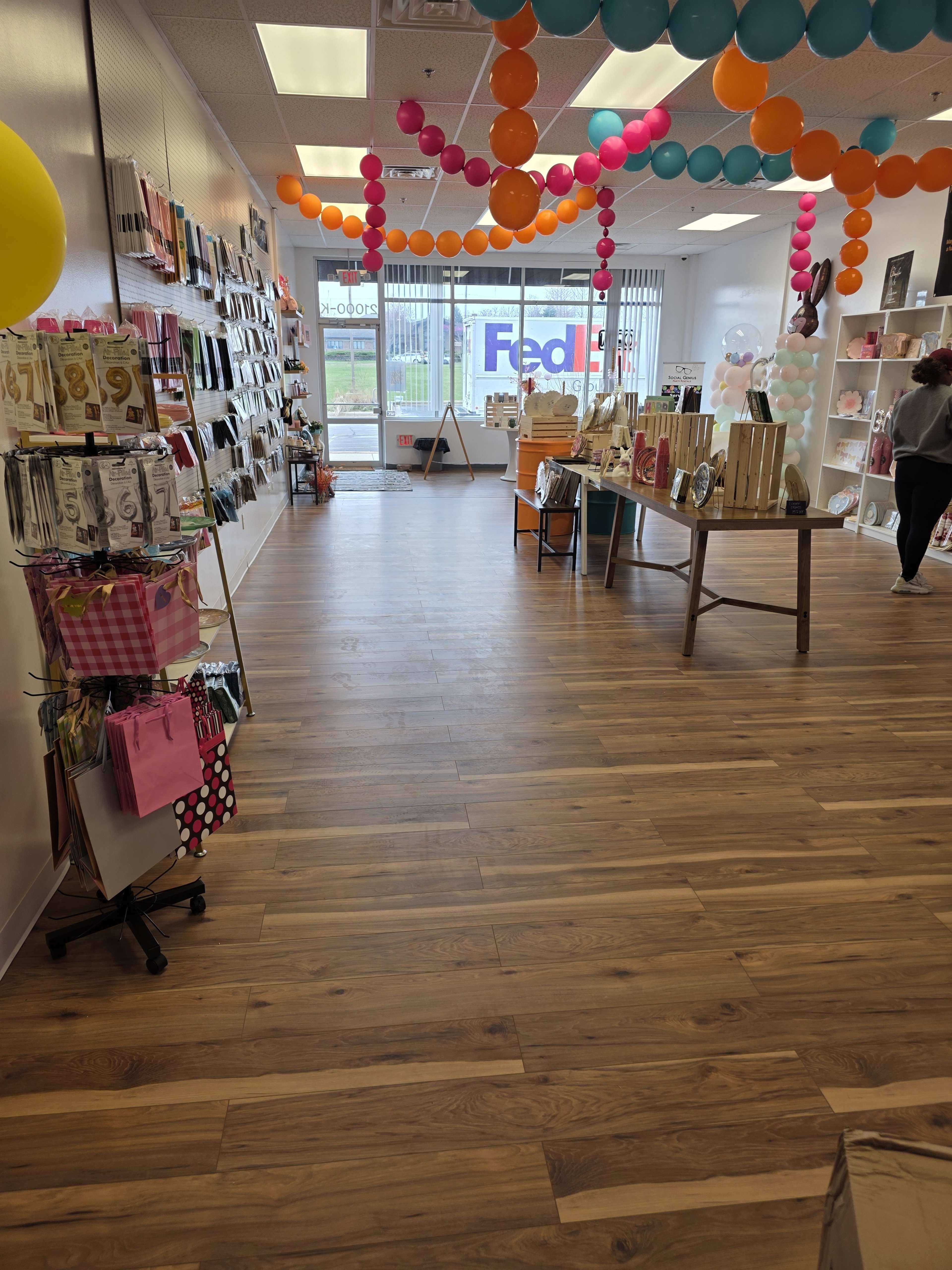 The image shows a brightly lit retail store with wooden flooring, decorated with colorful balloons and displaying various merchandise on shelves.