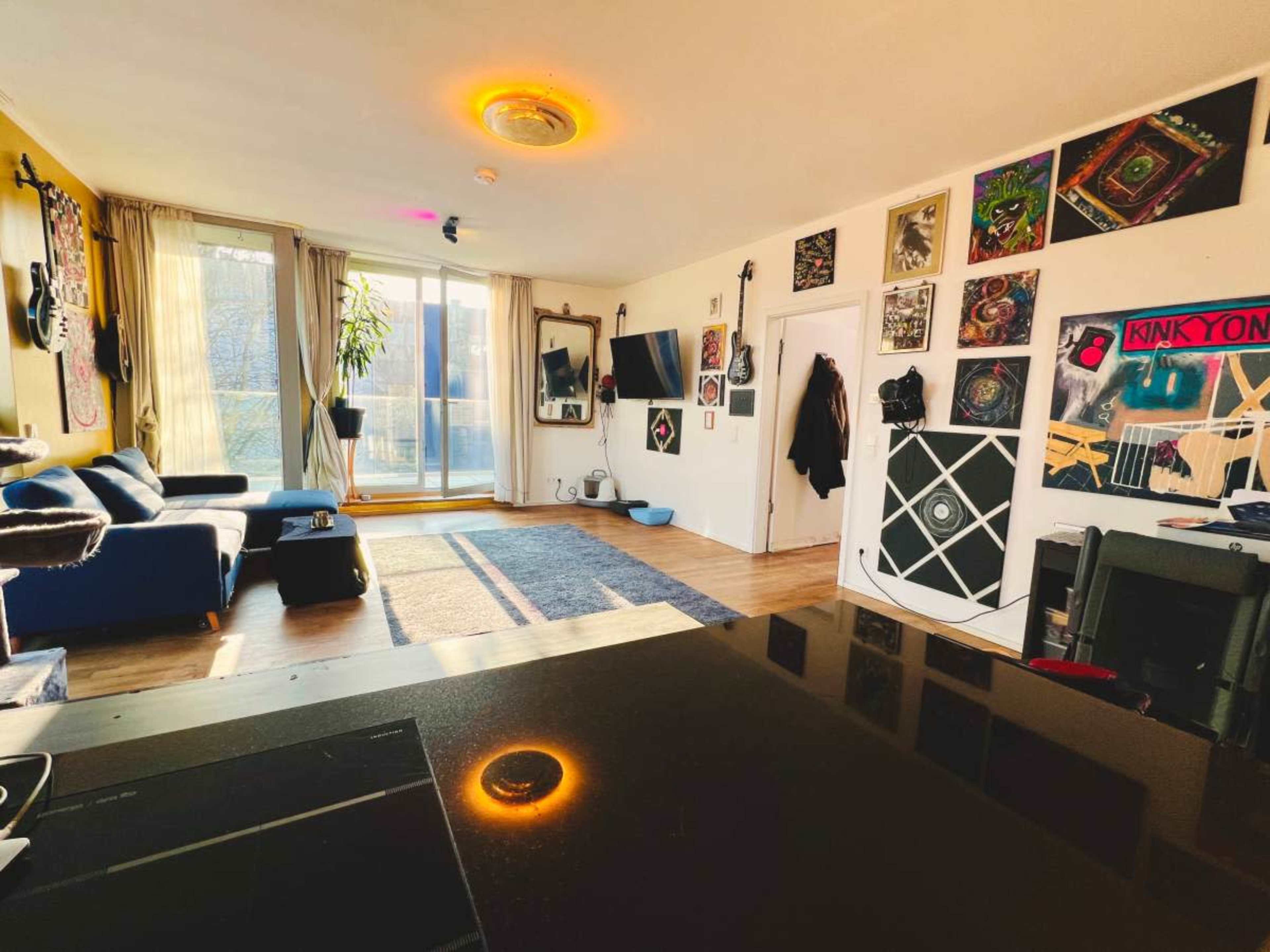 The image shows a brightly lit living room featuring a blue sofa, a large window, and various colorful artworks displayed on the walls.
