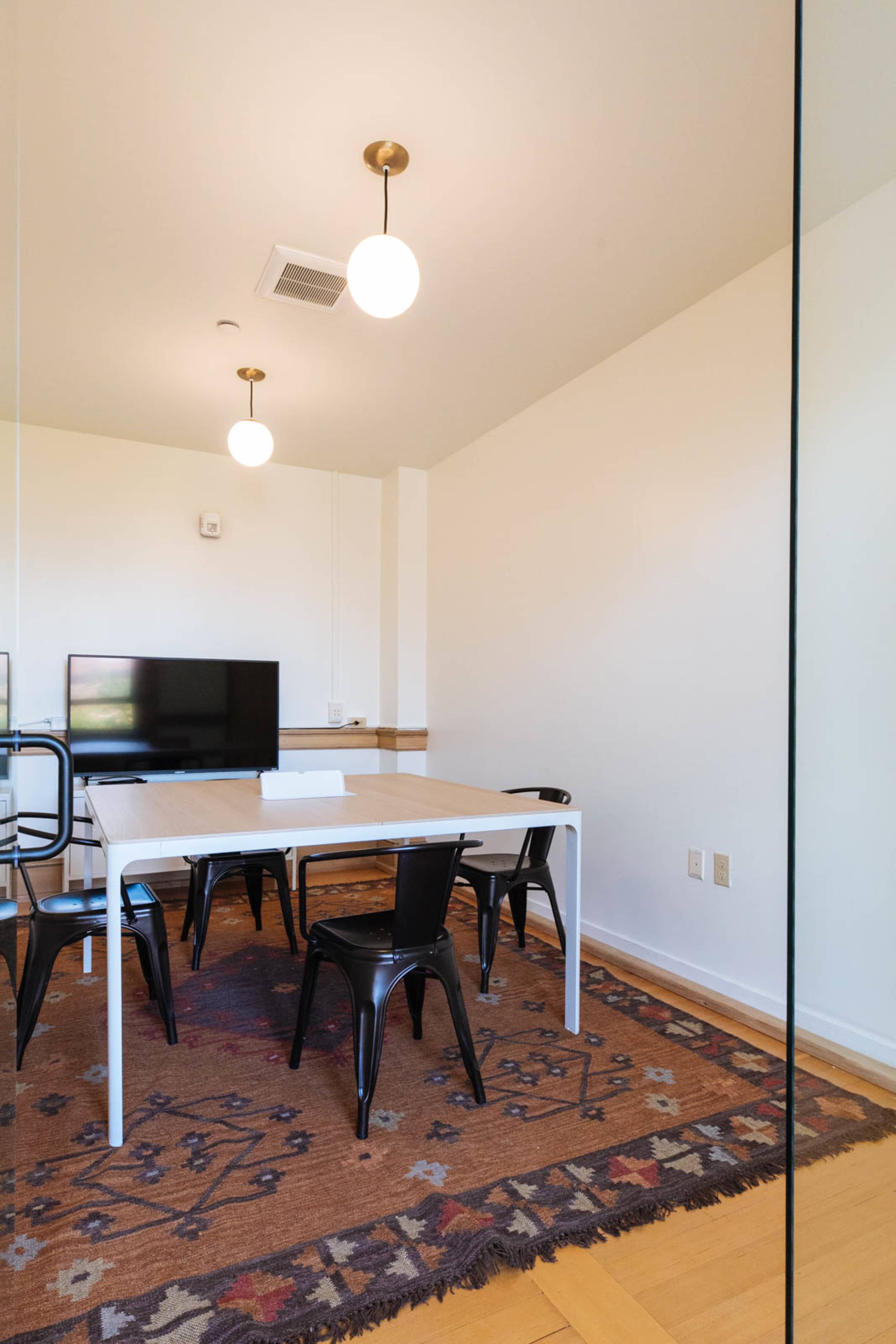 A modern conference room features a rectangular table surrounded by black chairs, with a television mounted on the wall and overhead pendant lights.