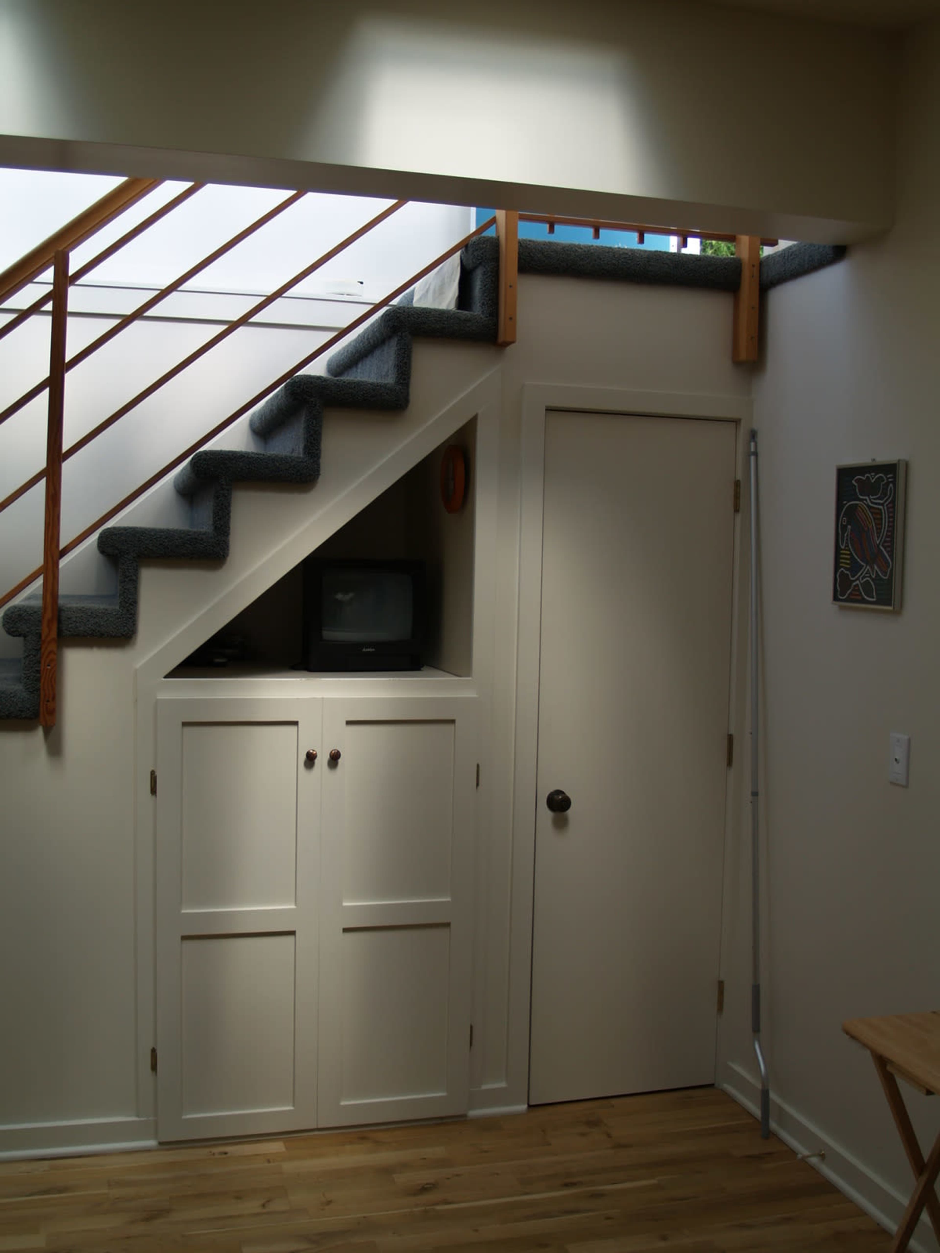 The image shows a staircase leading to an upper level, with a small cupboard and a door beneath the steps and a television placed on top.