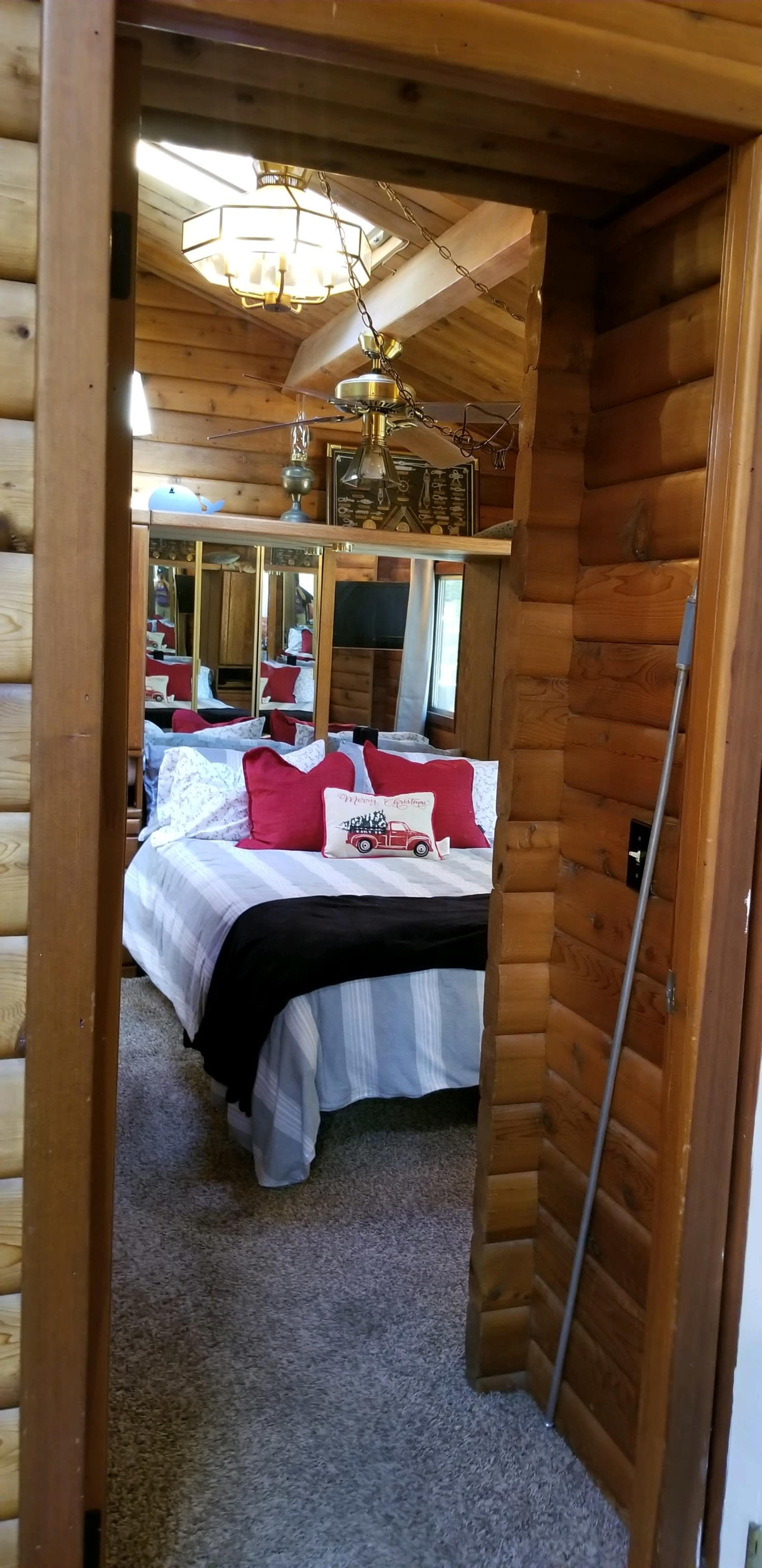 A cozy bedroom accessible through a wooden door, featuring a bed with red and white bedding, a decorative pillow, and a large mirror reflecting the room's interior.