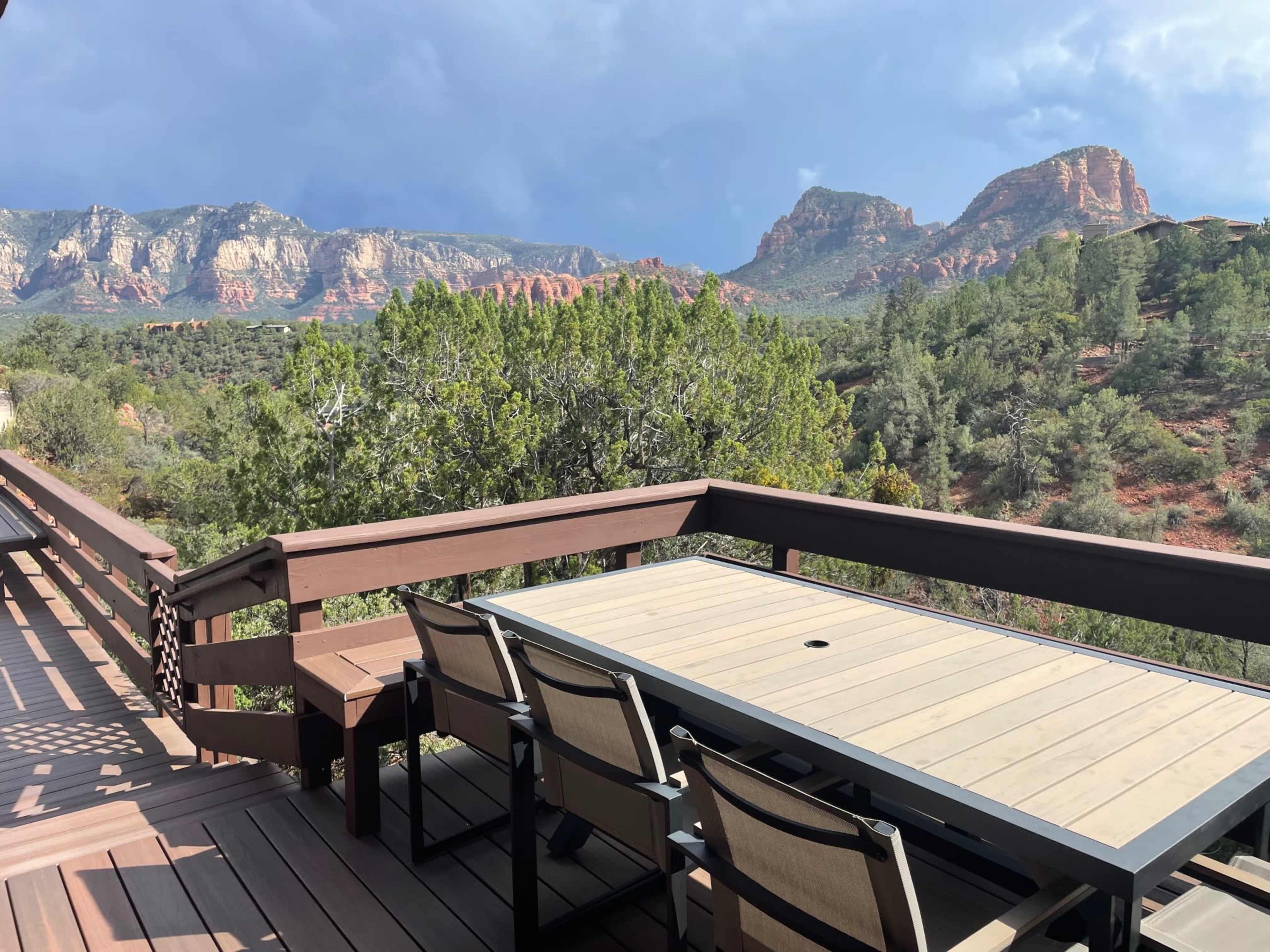 A wooden deck with a table and chairs overlooks a mountainous landscape with red rock formations and trees.